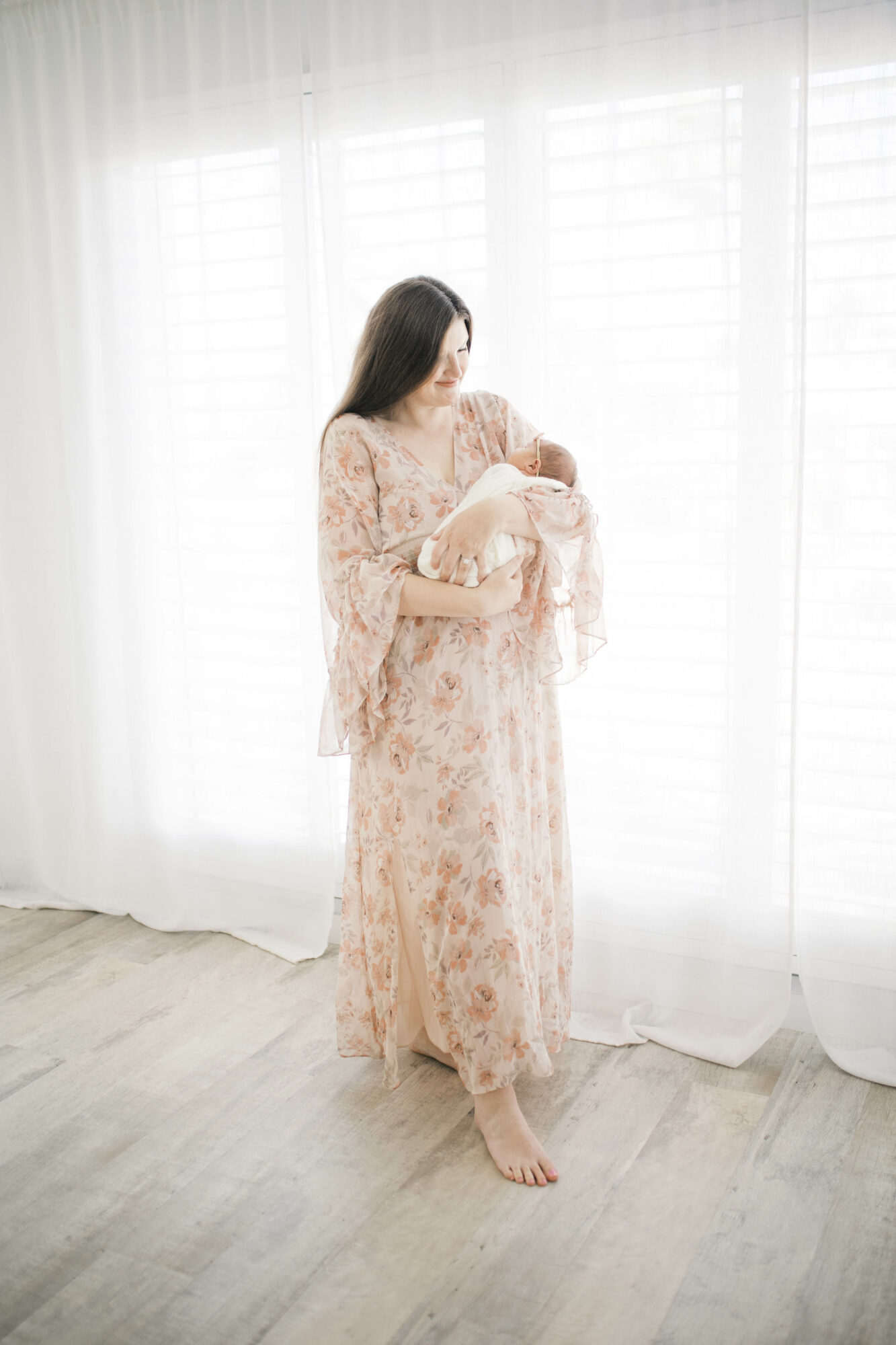 Woman in floral dress holding a baby, standing barefoot in front of sheer curtains, bright natural light, wooden floor