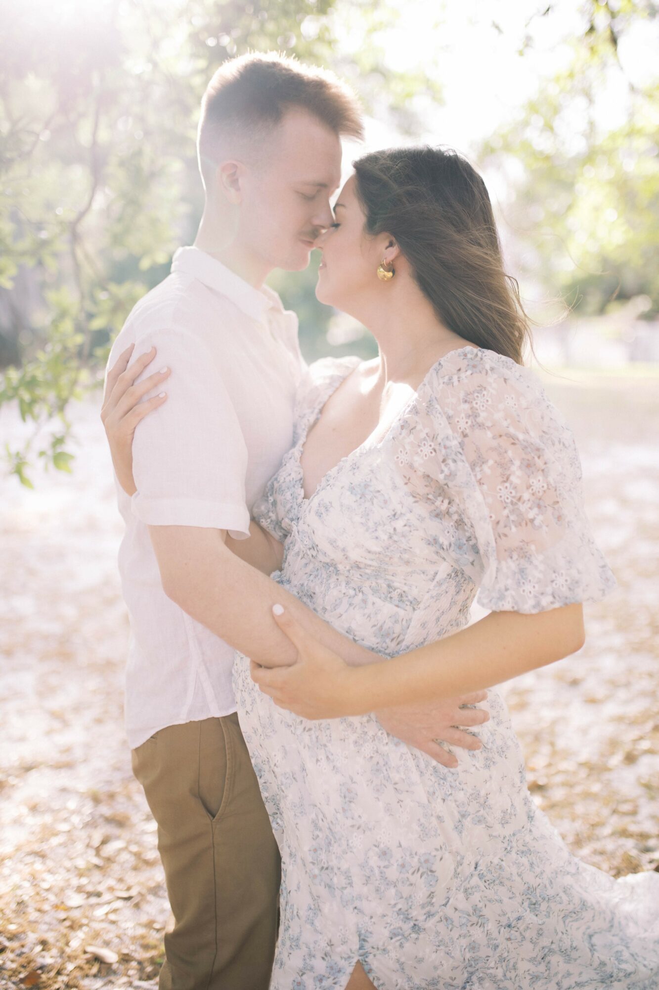 A couple stands close outdoors, gazing at each other, with trees and sunlight in the background.
