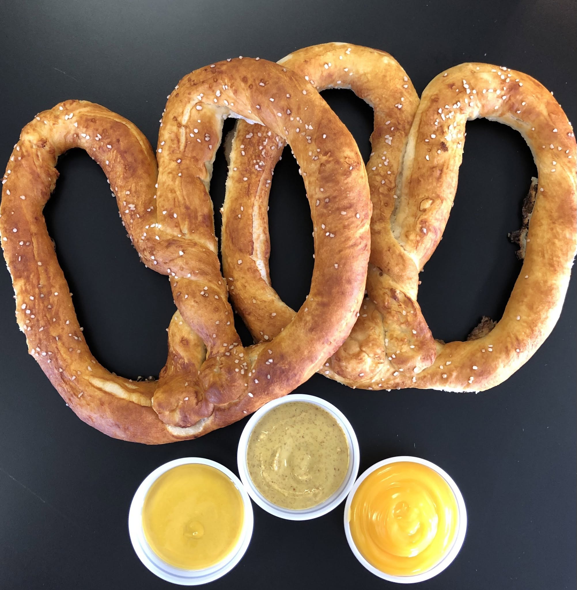 Three large pretzels with coarse salt, three small bowls of mustard and honey mustard, on a black surface.