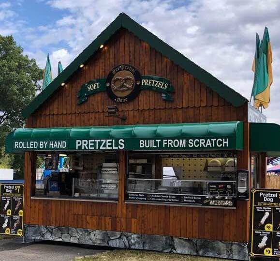 Wooden pretzel stand with green awning, signs advertising pretzels, and umbrellas in the background.