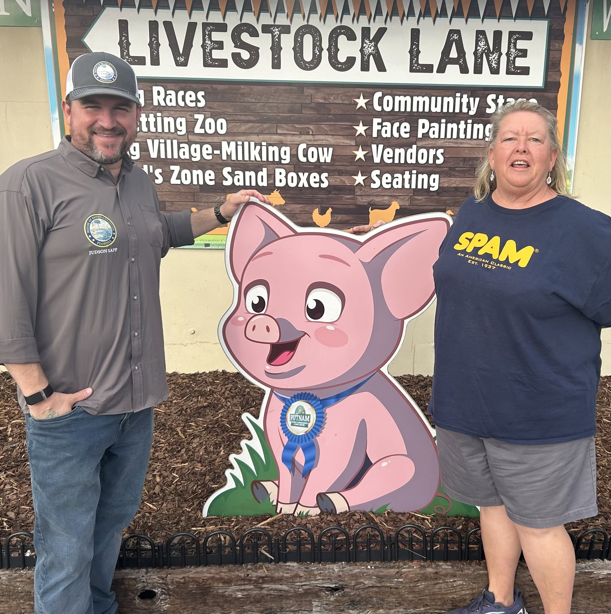 Man and woman standing beside a cartoon pig cutout at Livestock Lane event, smiling, with event sign in background.