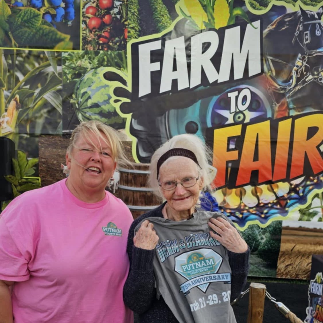 Two women smiling in front of a colorful farm fair banner, one wearing a pink shirt and the other a dark sweater, holding a bag.