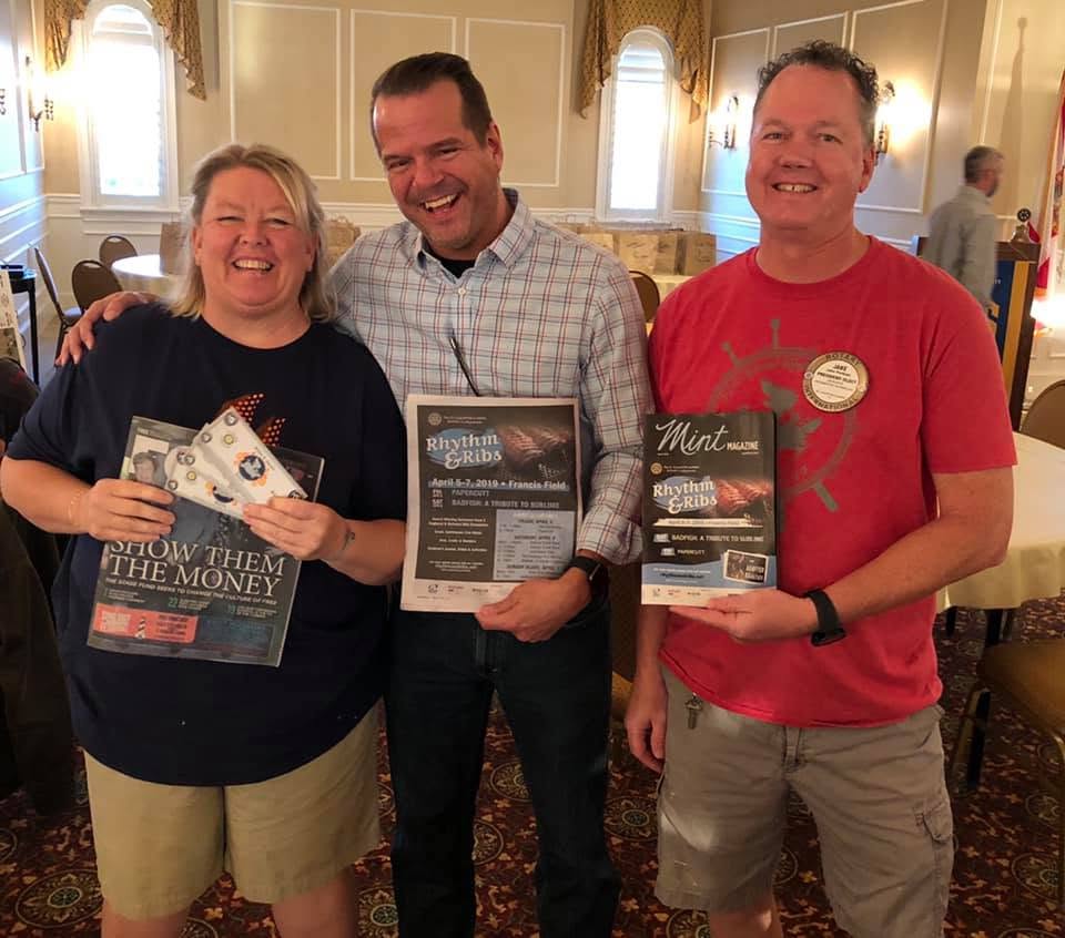 Three people smiling indoors, holding flyers, standing close together, with a woman on the left, a man in the middle, and another man on the right.