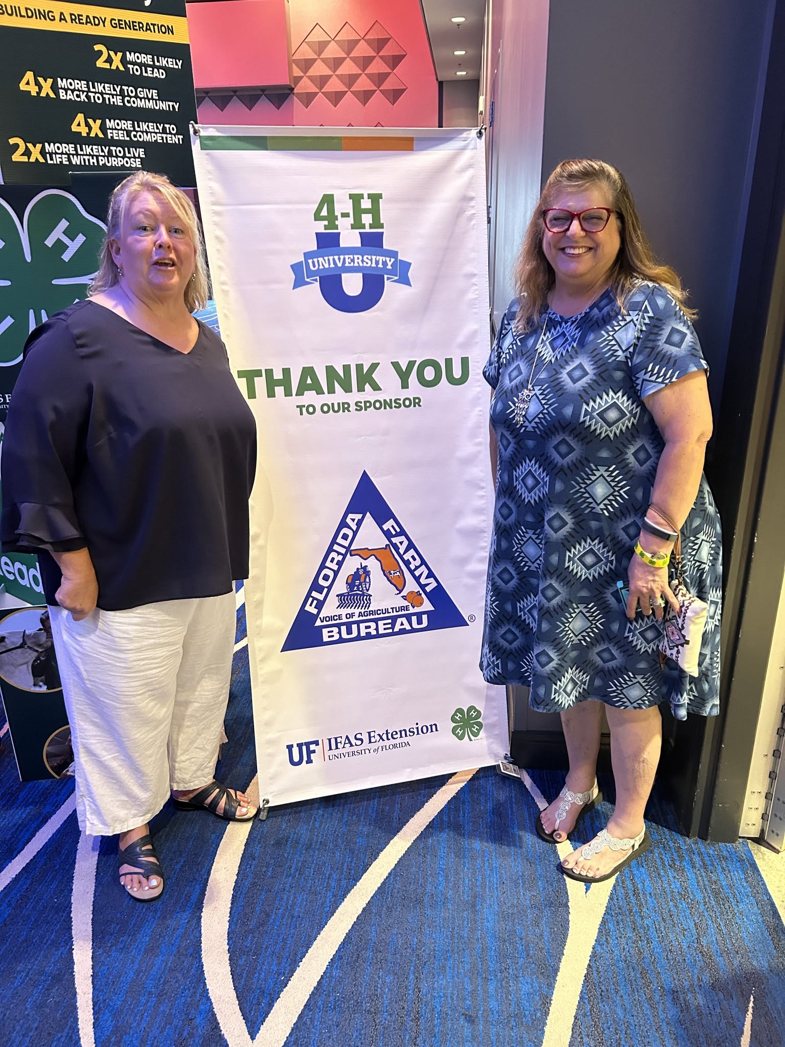 Two women stand on a blue patterned carpet, smiling, next to a thank you banner for a sponsor, in an indoor event space.