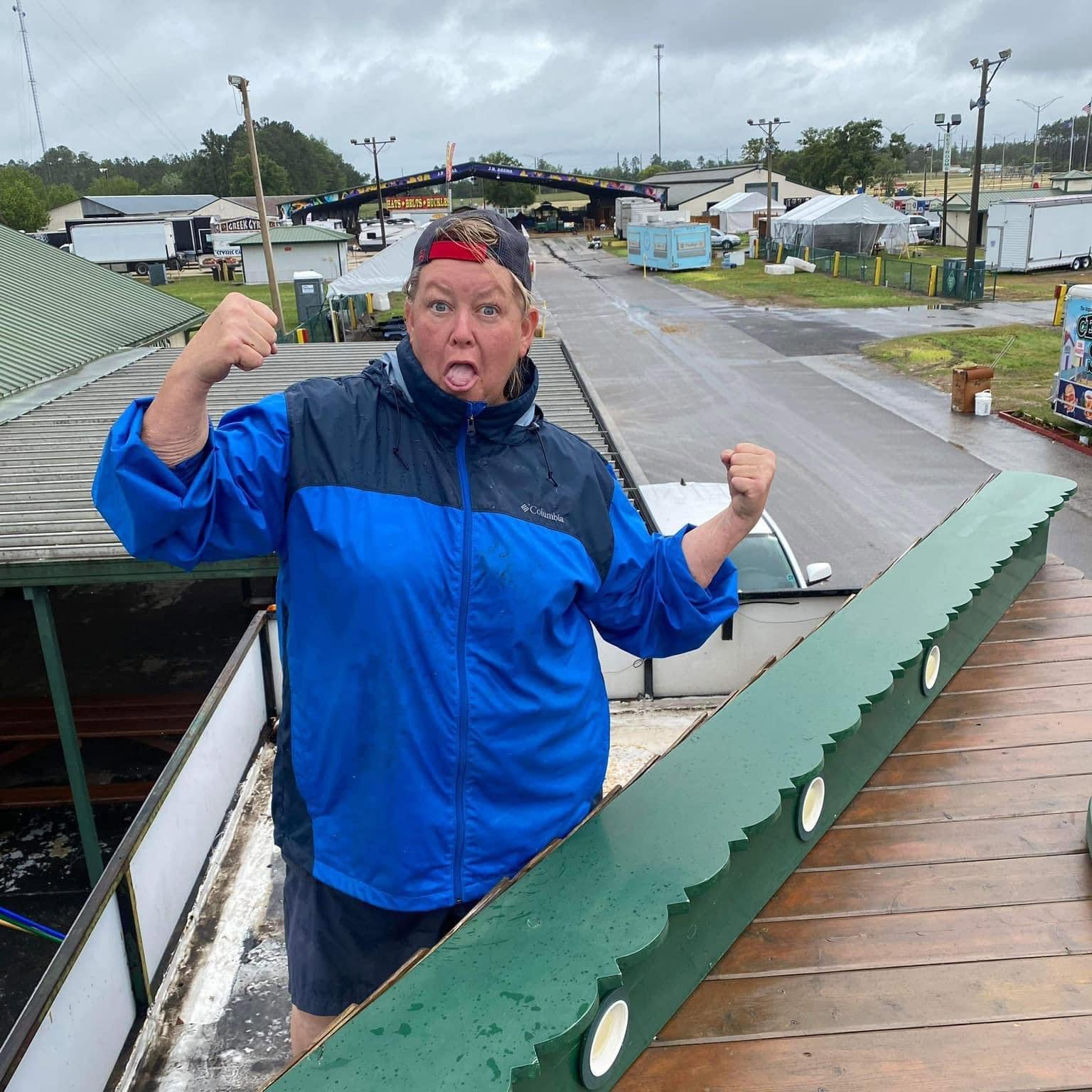 Person wearing a blue jacket making a fist with both hands, standing outdoors near a wooden railing, with a surprised expression.