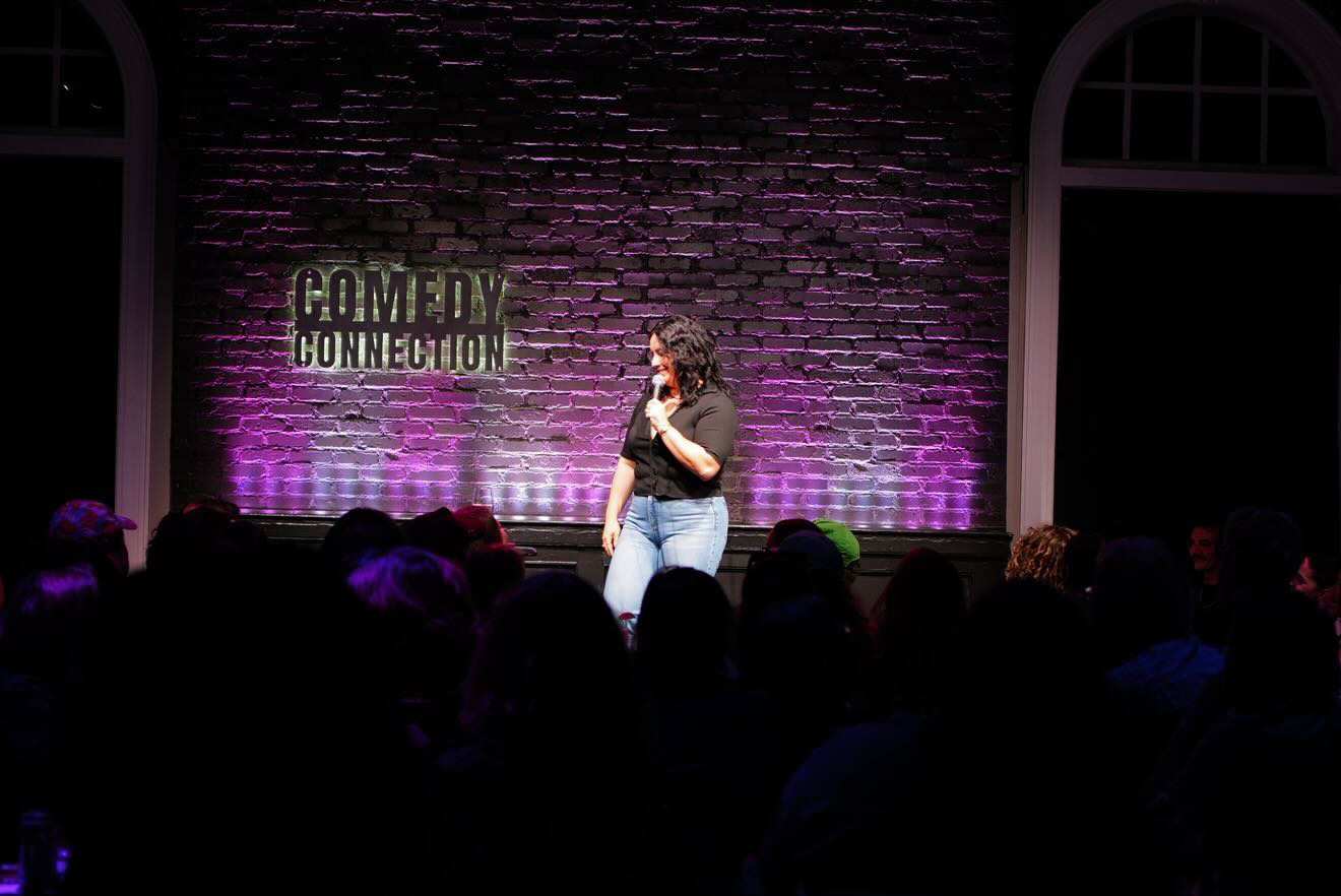 Person standing on stage with a microphone, audience in foreground, brick wall background with purple lighting, and a sign reading 'Comedy Connection'.