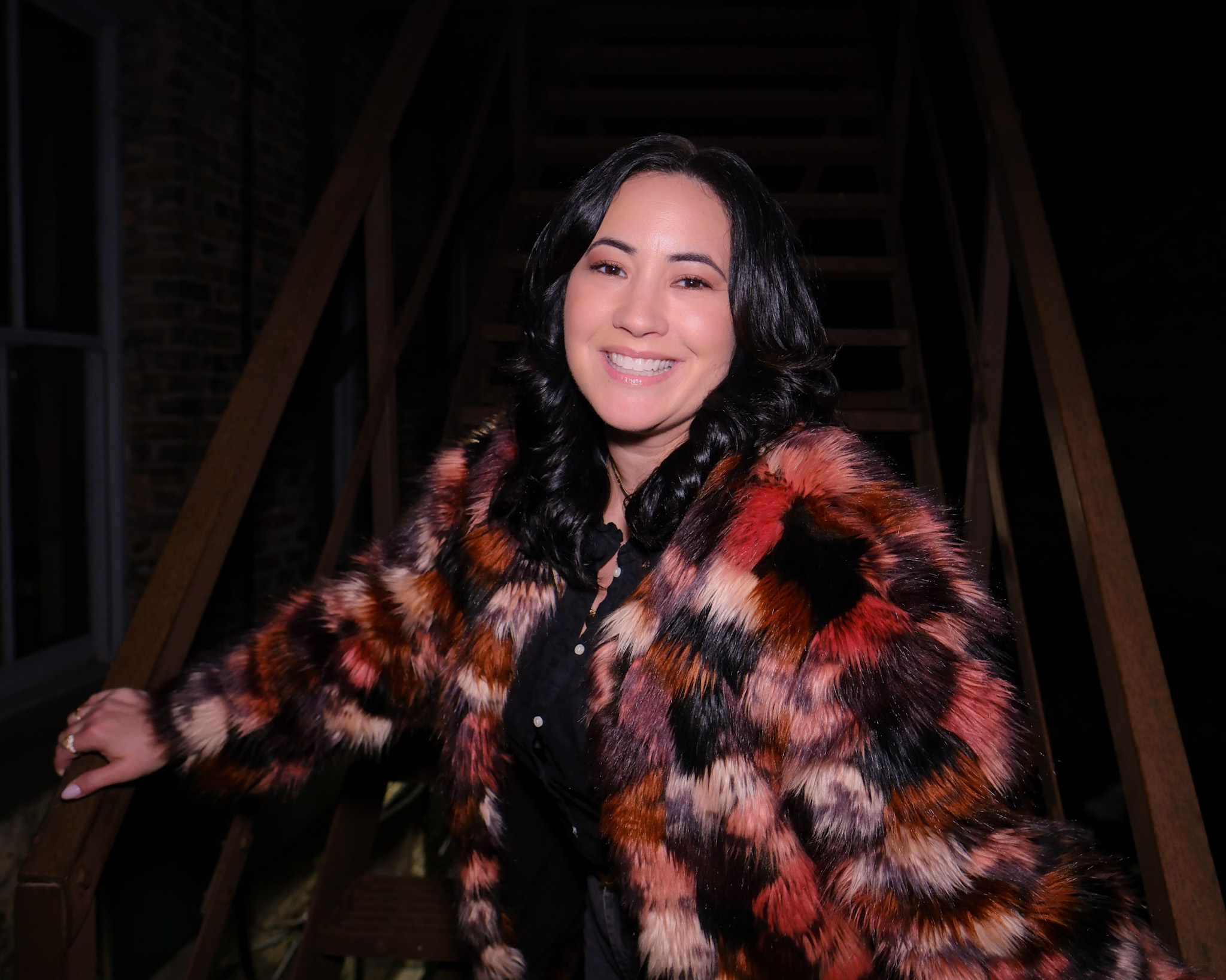 Woman with dark hair smiling, wearing a colorful fur coat, standing on stairs in a dark setting.