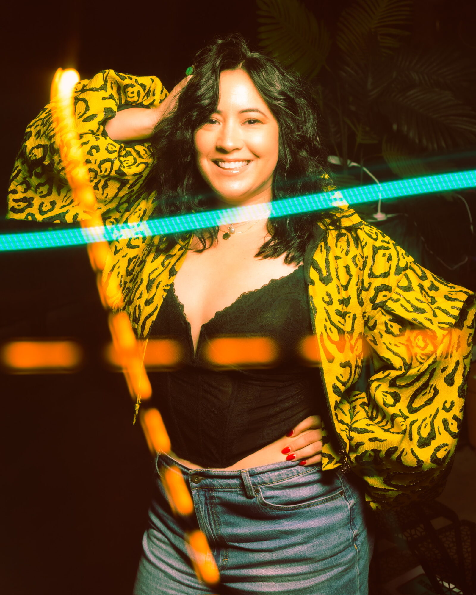 Smiling woman with wavy hair wearing a yellow patterned jacket and black top, standing in a dark setting with colorful lights.