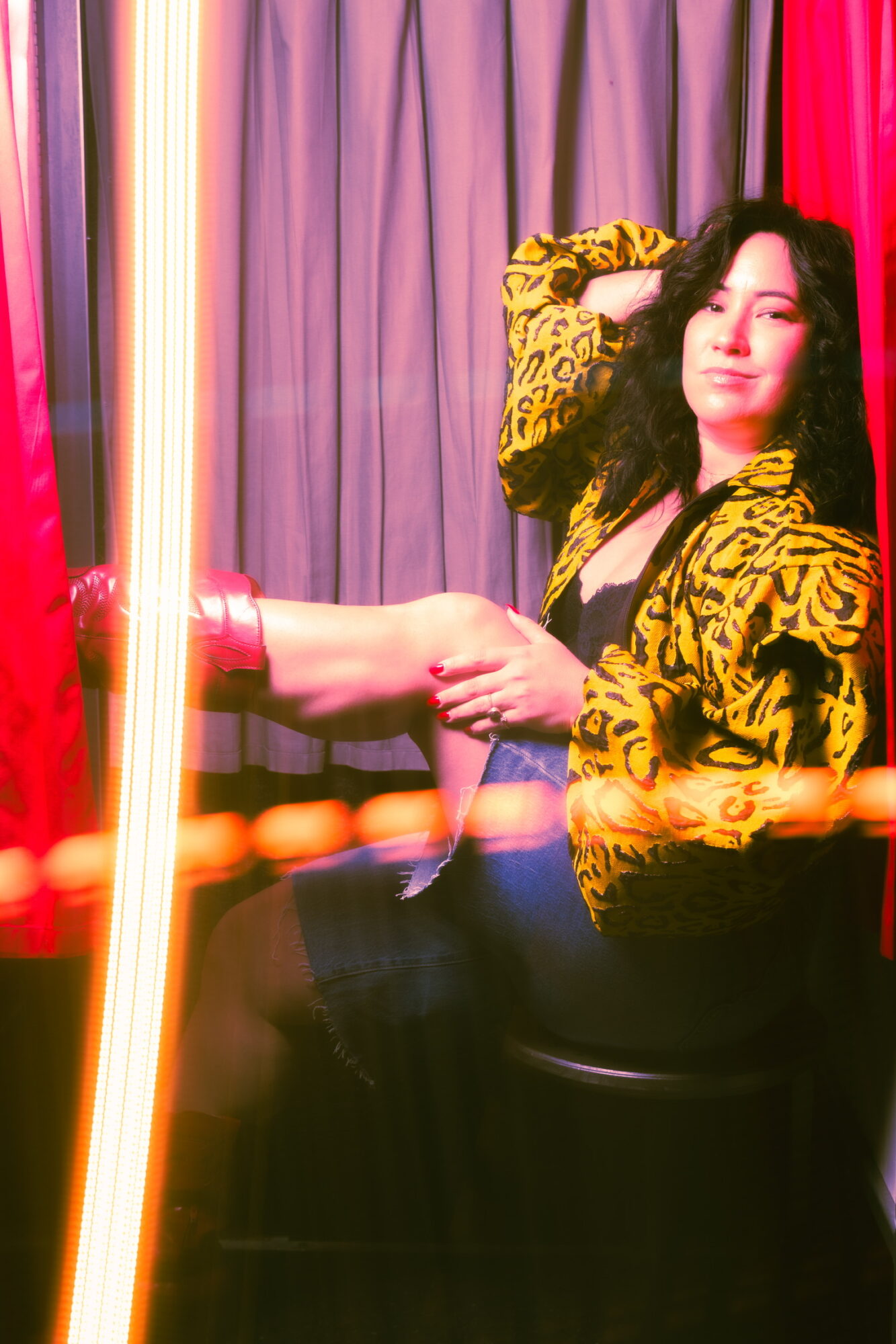 Woman with long dark hair sitting with legs crossed, wearing a yellow and black patterned jacket, in front of colorful curtains.