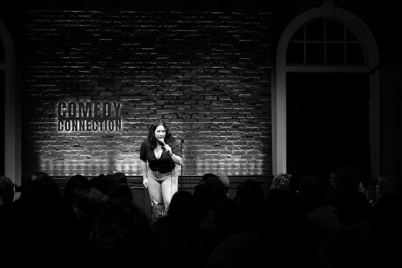 Woman speaking on stage with a brick wall background and audience in foreground.