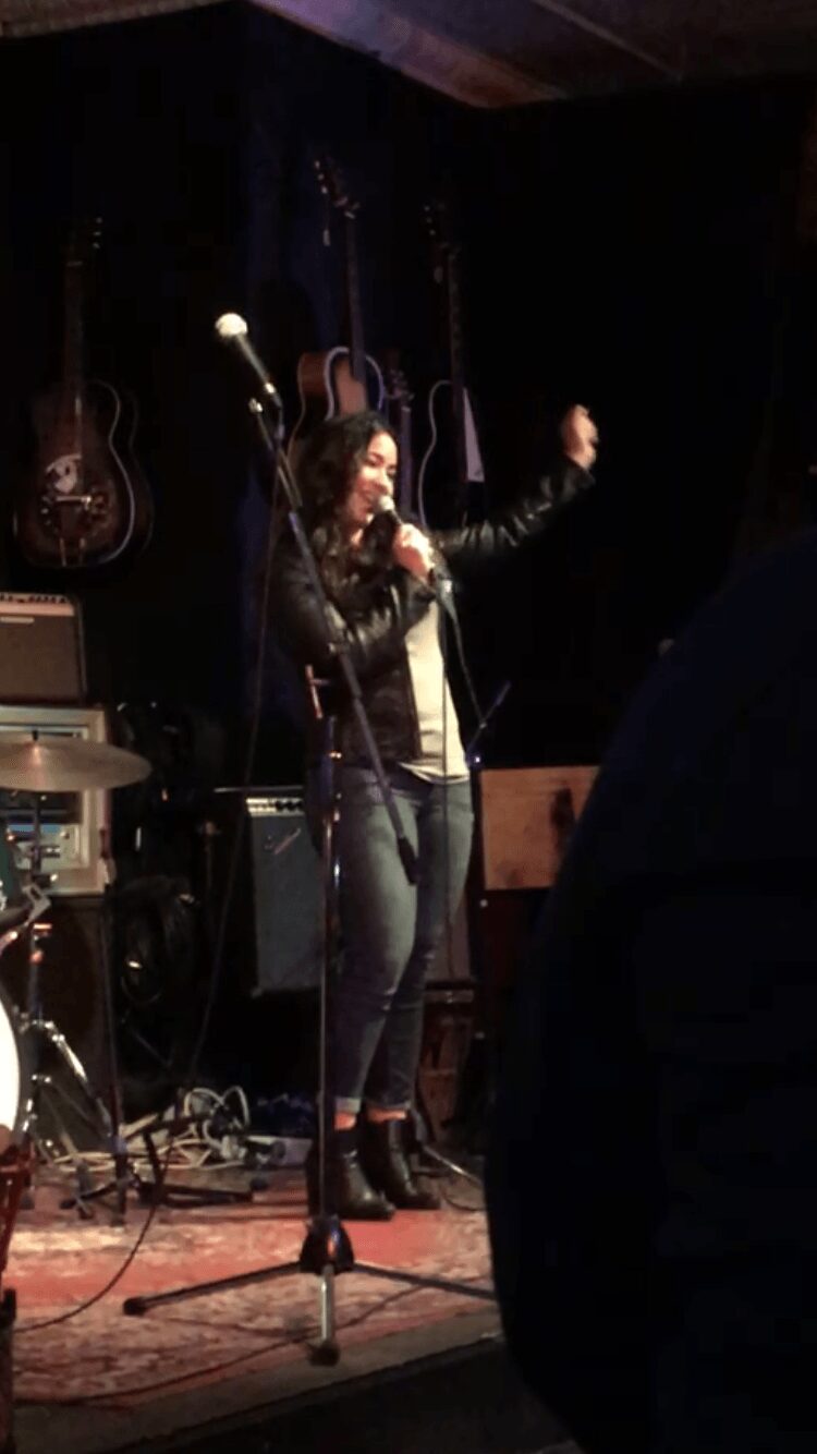 A woman singing on stage with a microphone, surrounded by musical instruments and equipment, in a dark venue.