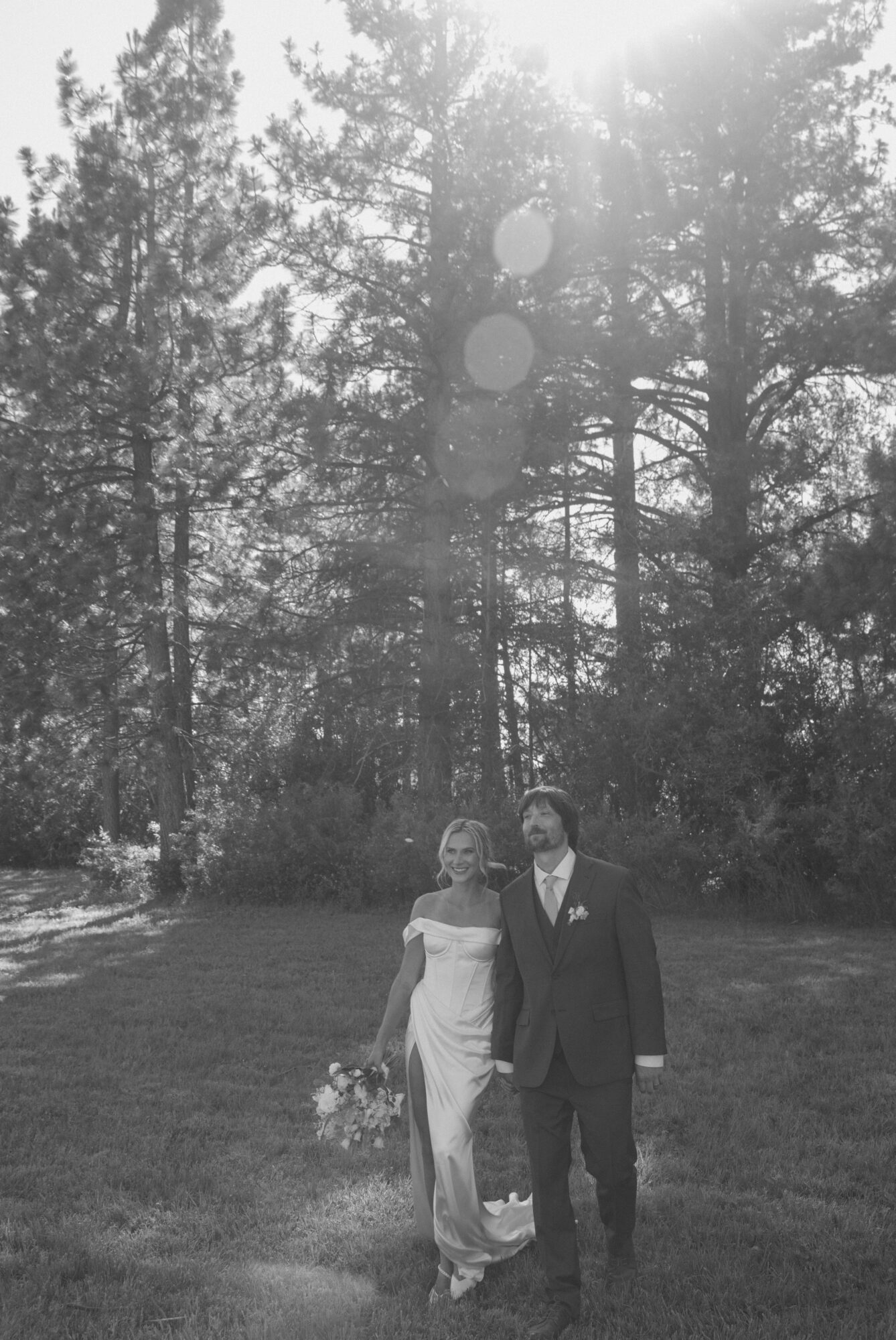 Two people in formal attire standing outdoors on grass with trees in background, sunlight shining through trees.