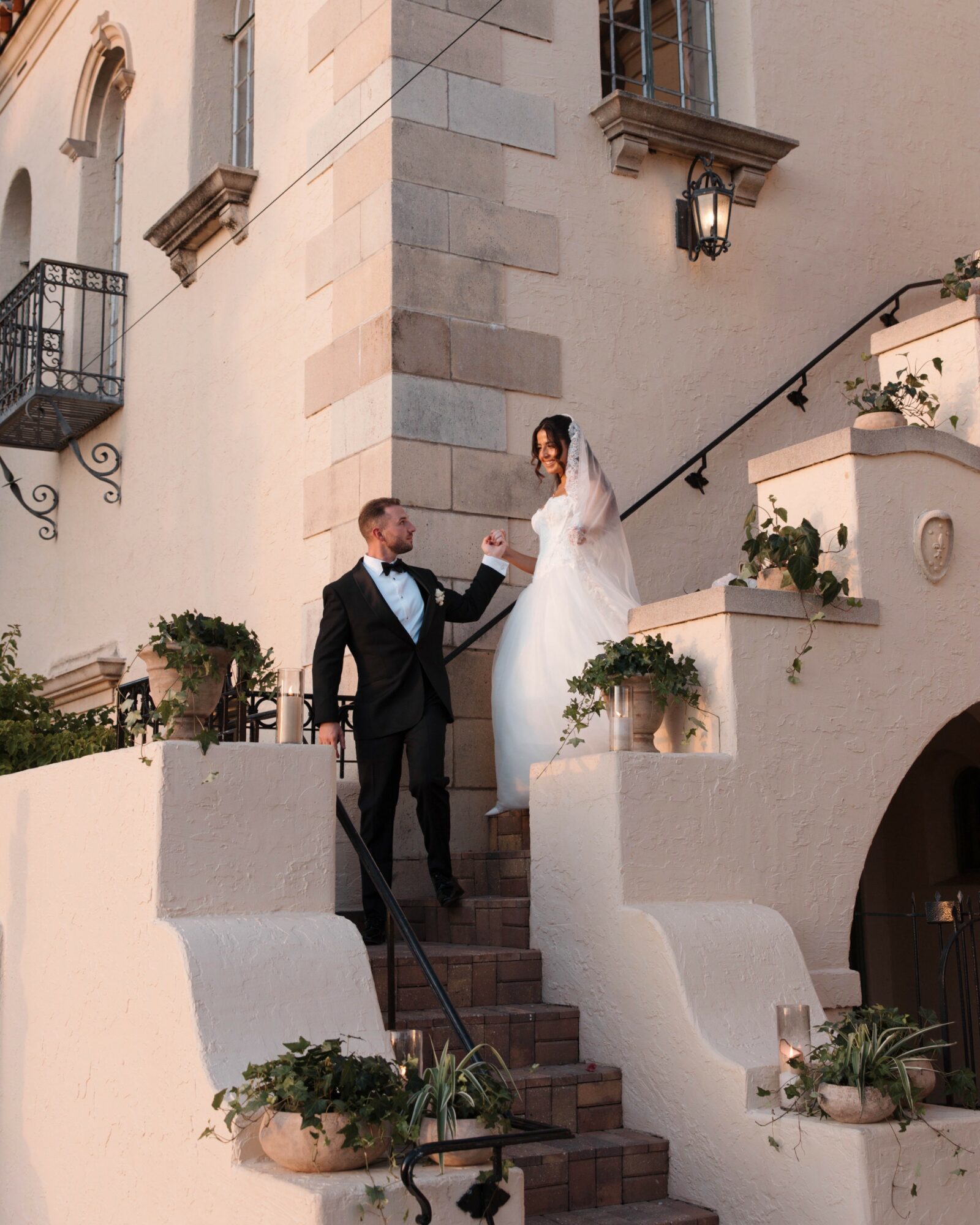 A bride and groom on stairs outside a building, bride in white gown, groom in black suit, holding hands.