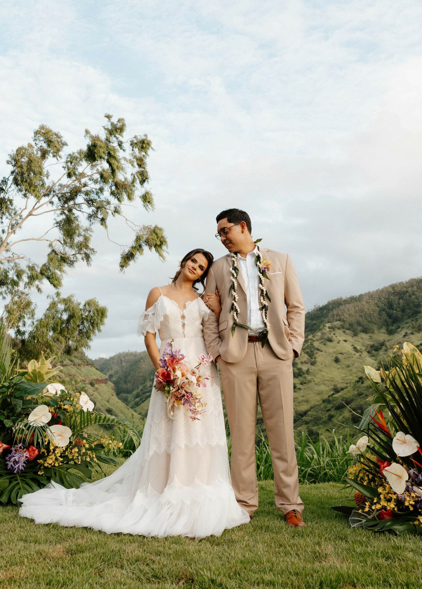Bride and groom standing outdoors with mountains and trees, smiling, surrounded by flowers, on a grassy field.