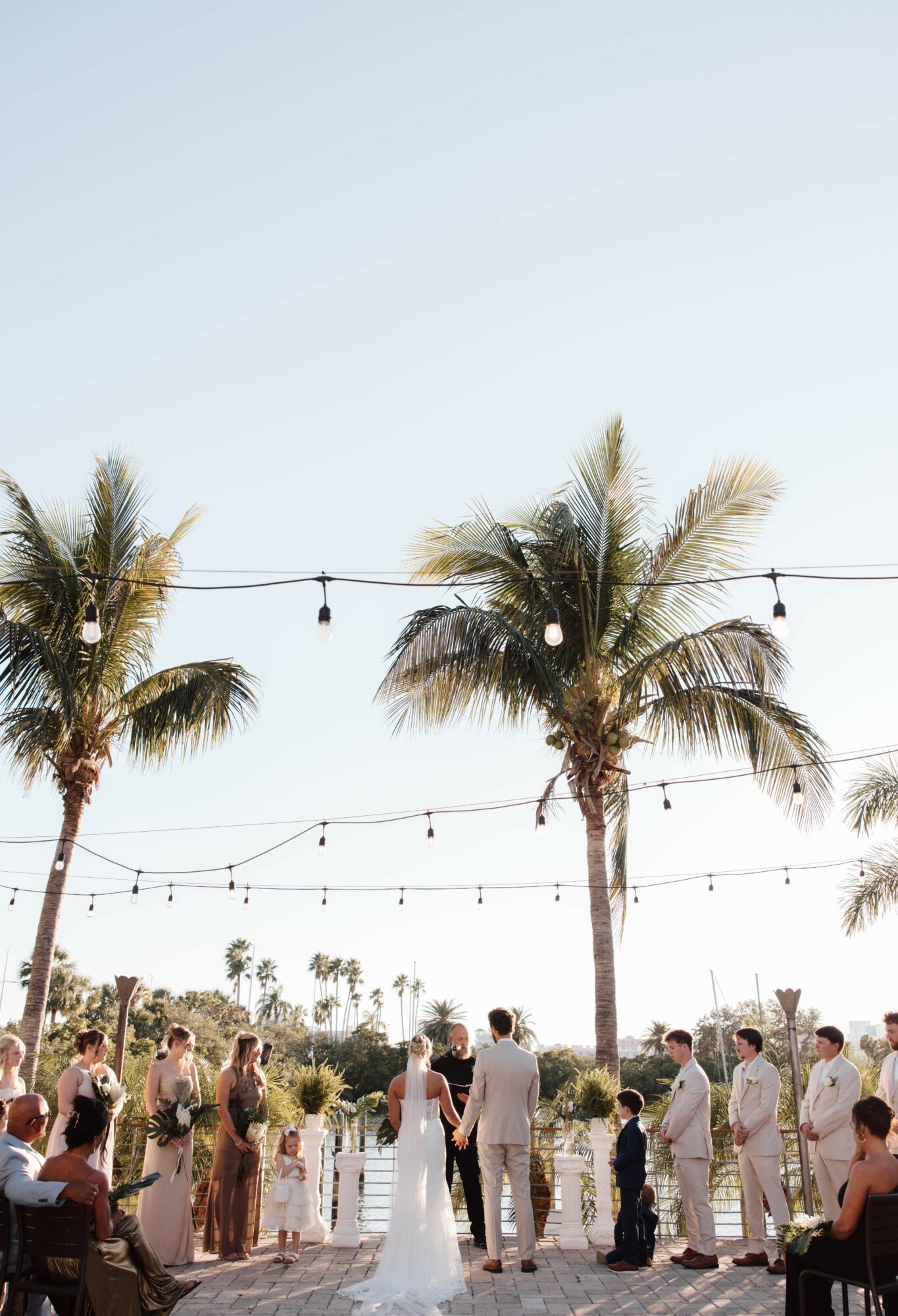 Outdoor wedding ceremony with palm trees, string lights, and guests, under a clear sky.