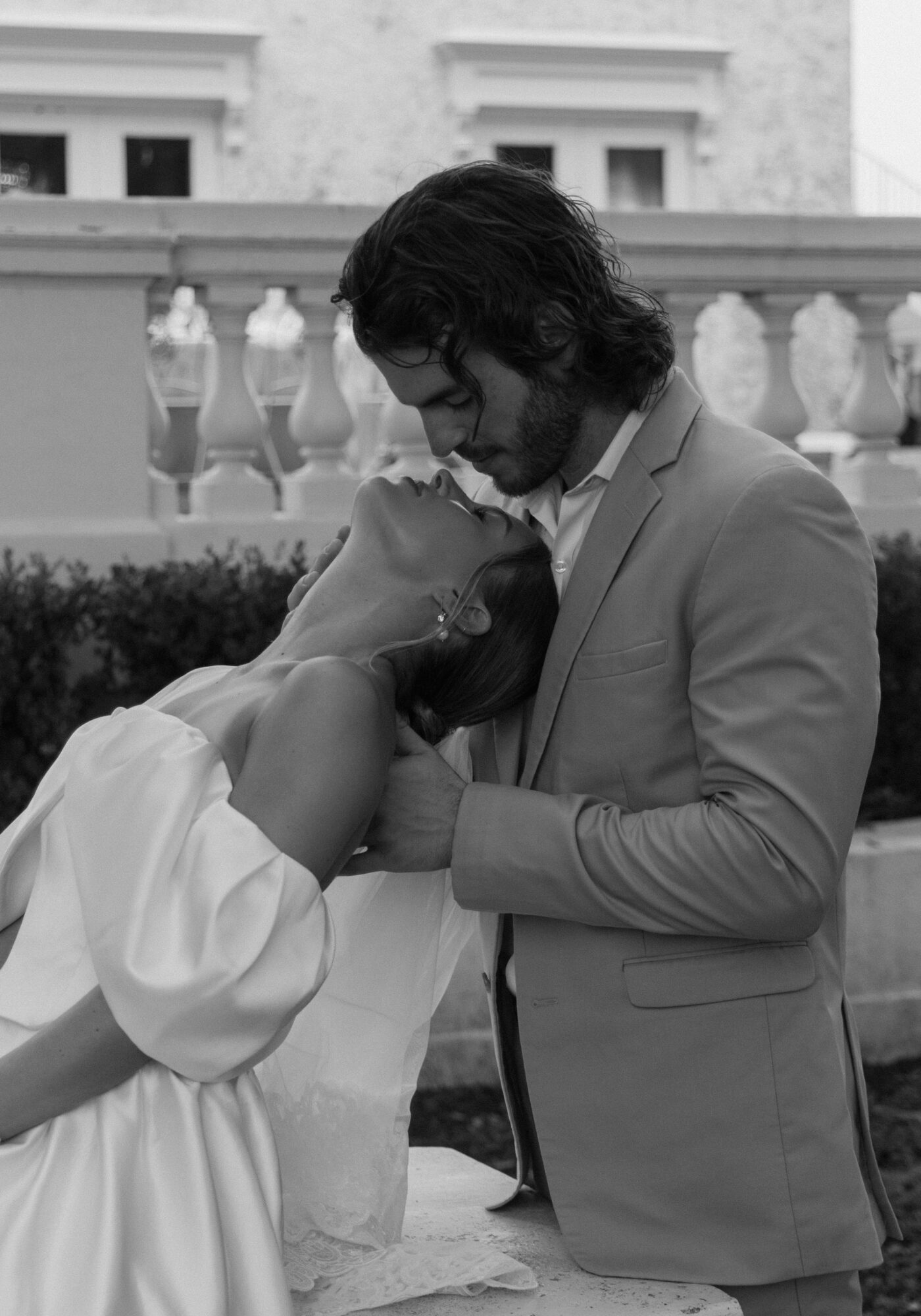 A man and woman share an intimate moment outdoors, with the woman leaning on the man's chest and the man holding her gently.
