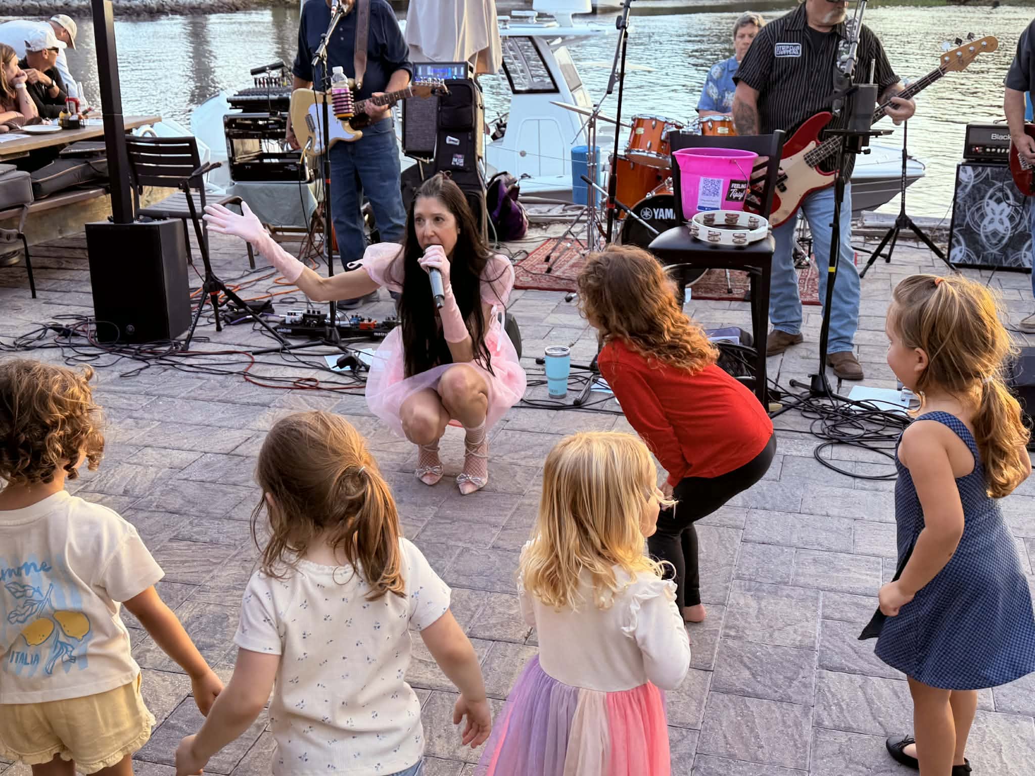 Group of children watching a singer perform outdoors near water, with musicians playing instruments in the background.