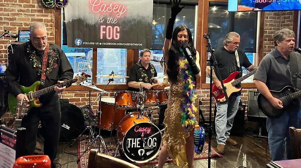 Band performing live with four musicians, one singing, in a brick-walled venue with a sign reading 'Casey & the Fog'.