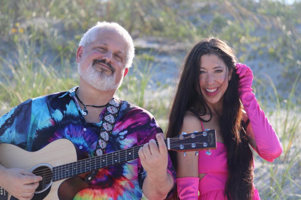 Two people outdoors, one playing guitar and smiling, the other smiling and touching her hair, with greenery in background.