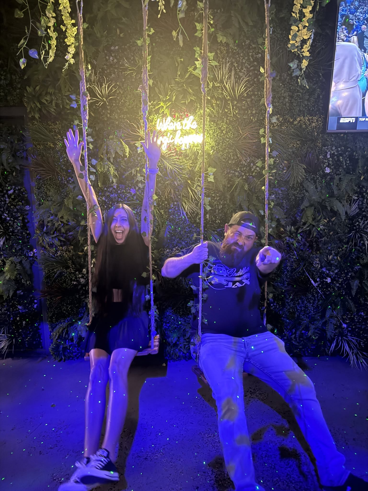 Two people sitting on swings outdoors at night, surrounded by trees and illuminated by colorful lights.