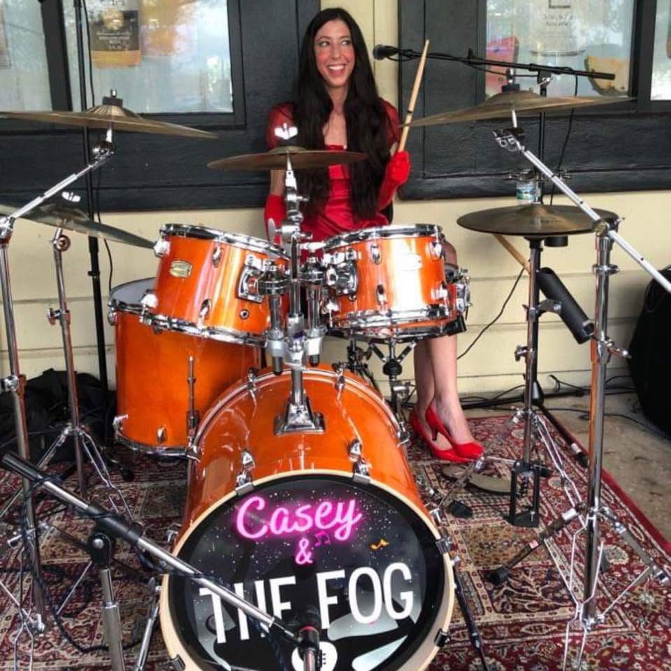 Woman sitting behind a drum set with a sign reading 'Casey & The Fog' in front, smiling, in a room with large windows.