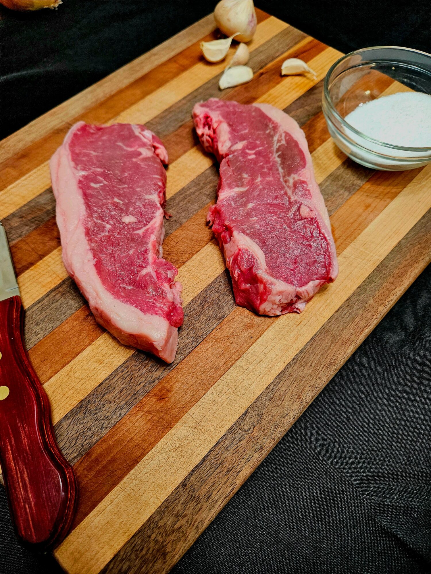 Two raw beef steaks on a wooden cutting board with garlic and a bowl of seasoning nearby.