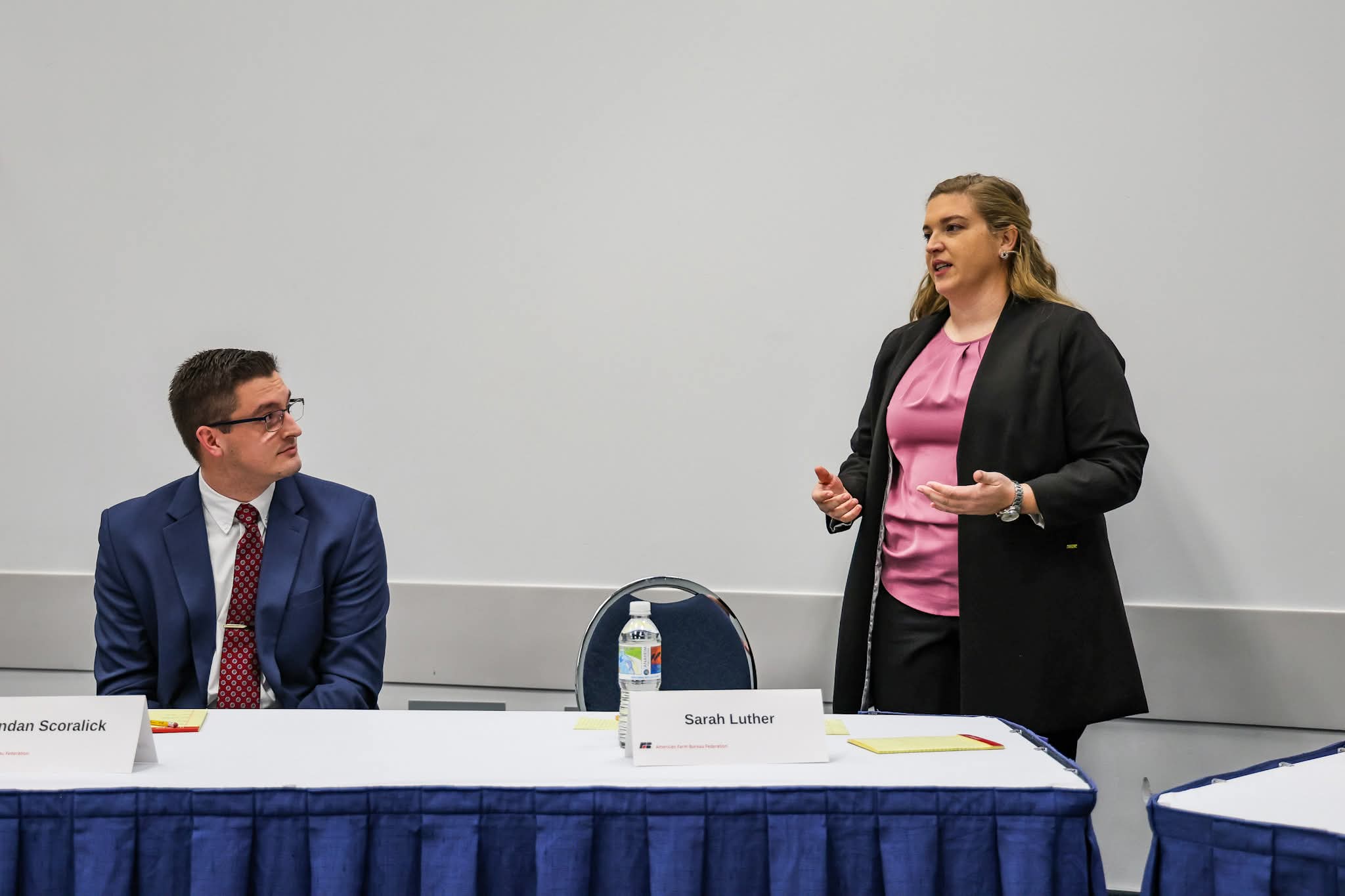 Two women in a meeting room, one standing and speaking, the other sitting and listening, with a table and name tags.