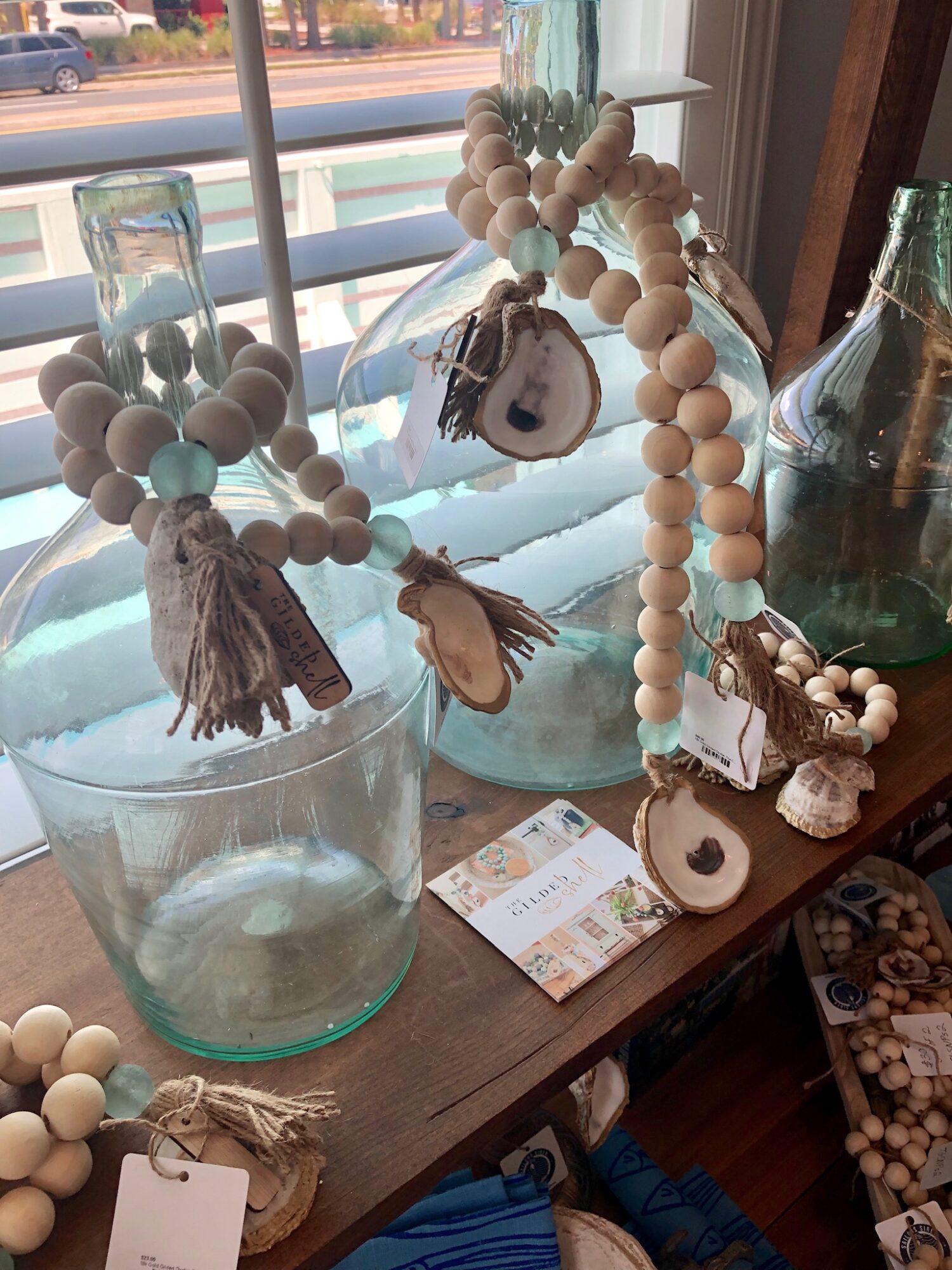Decorative beaded ornaments with tassels and tags on a wooden surface near a window, with glass bottles and a card nearby.