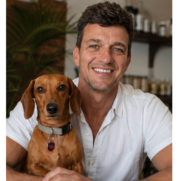 Man smiling with a small brown dog, both looking at the camera, indoors with shelves in background.