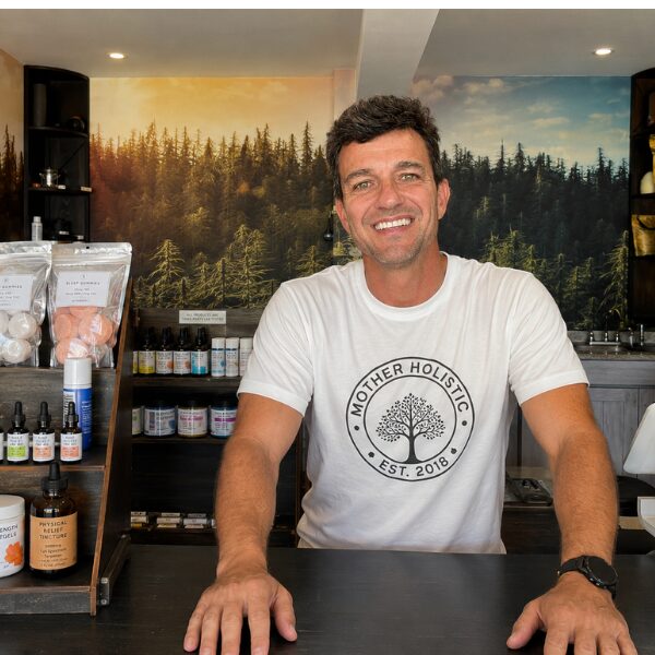 Smiling man standing behind a counter in a store with shelves of products and a forest landscape mural in the background.