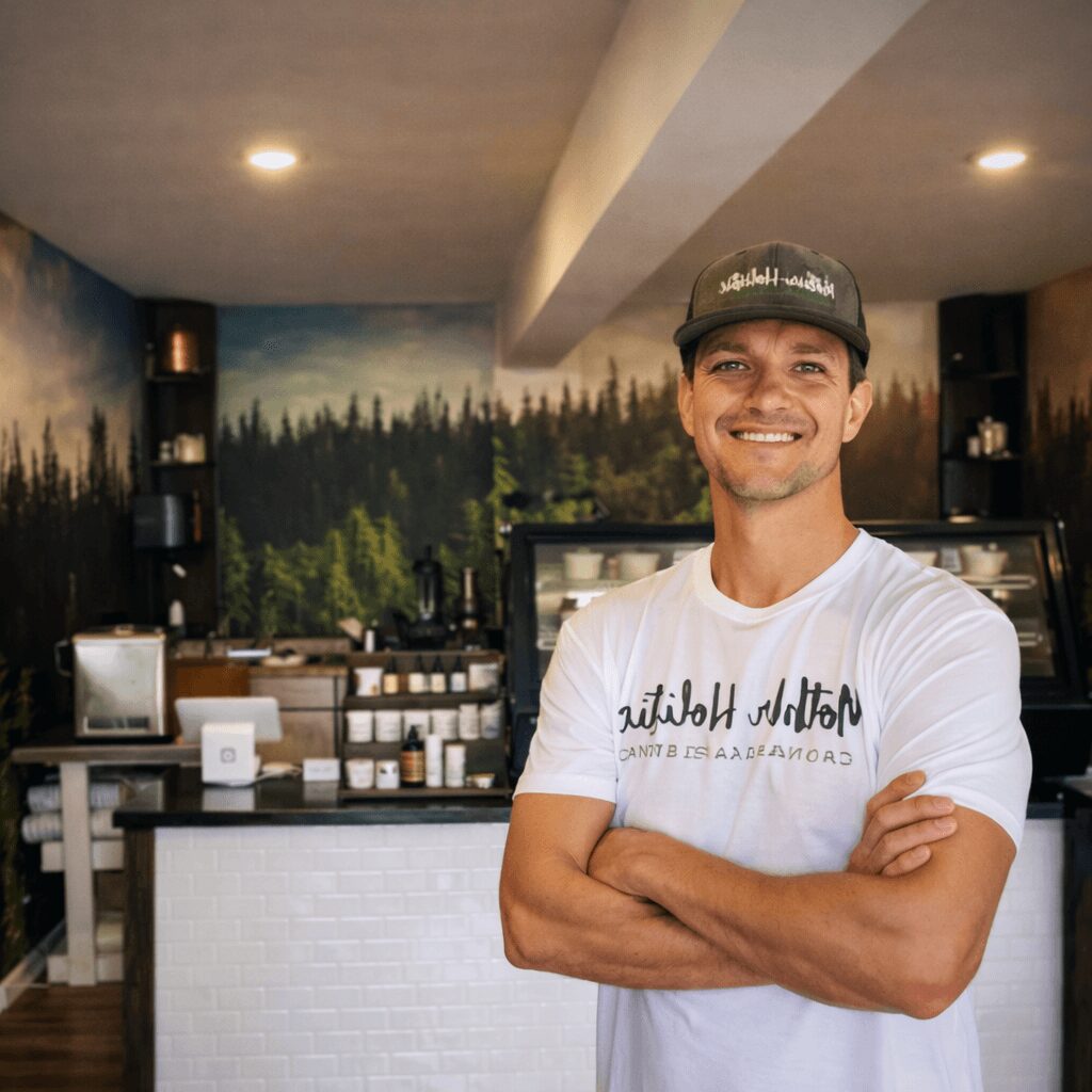 Smiling man with arms crossed in a coffee shop, background features a forest mural and coffee equipment.