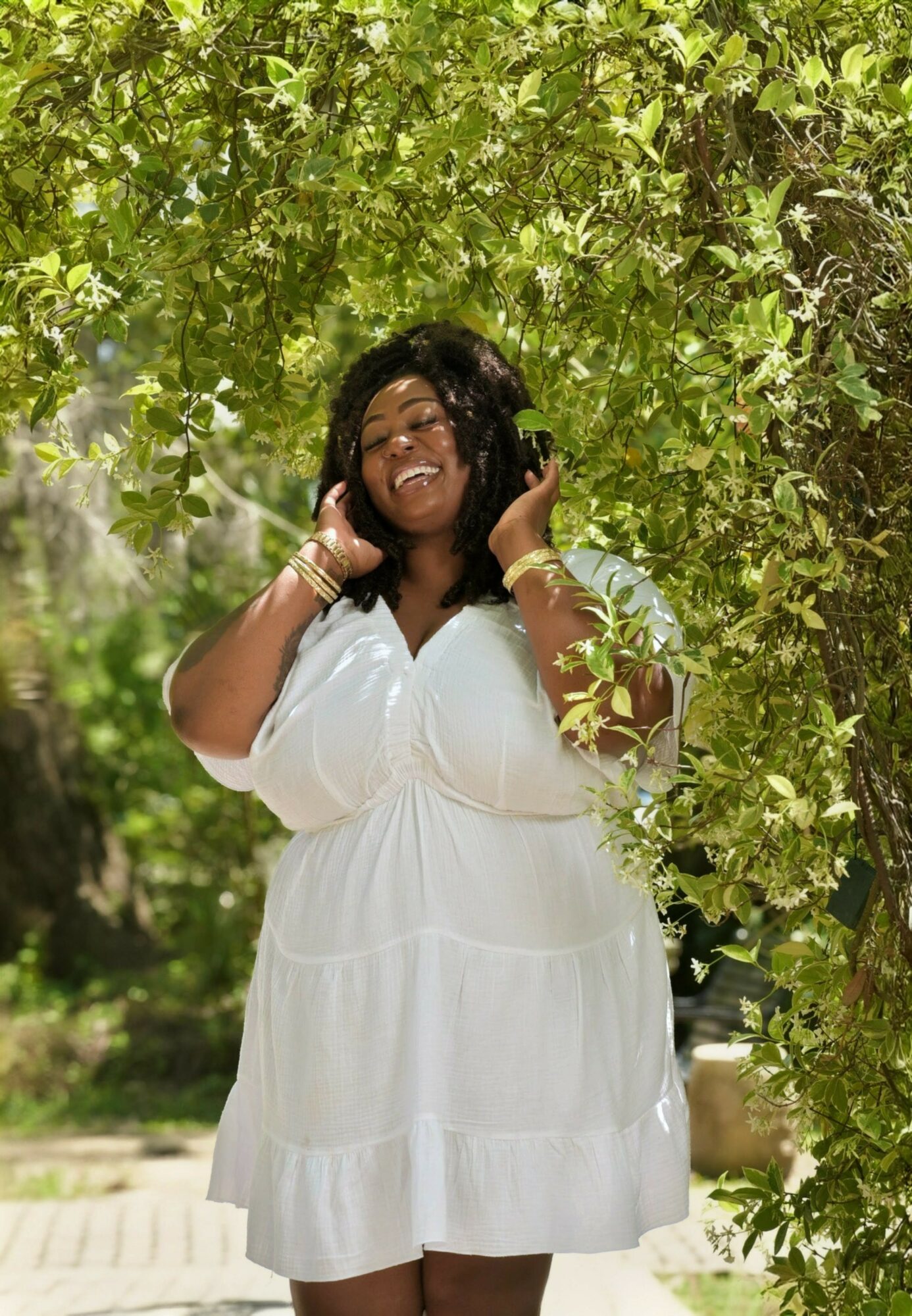 Smiling woman in white dress standing under green leafy tree, holding her hair, outdoors.