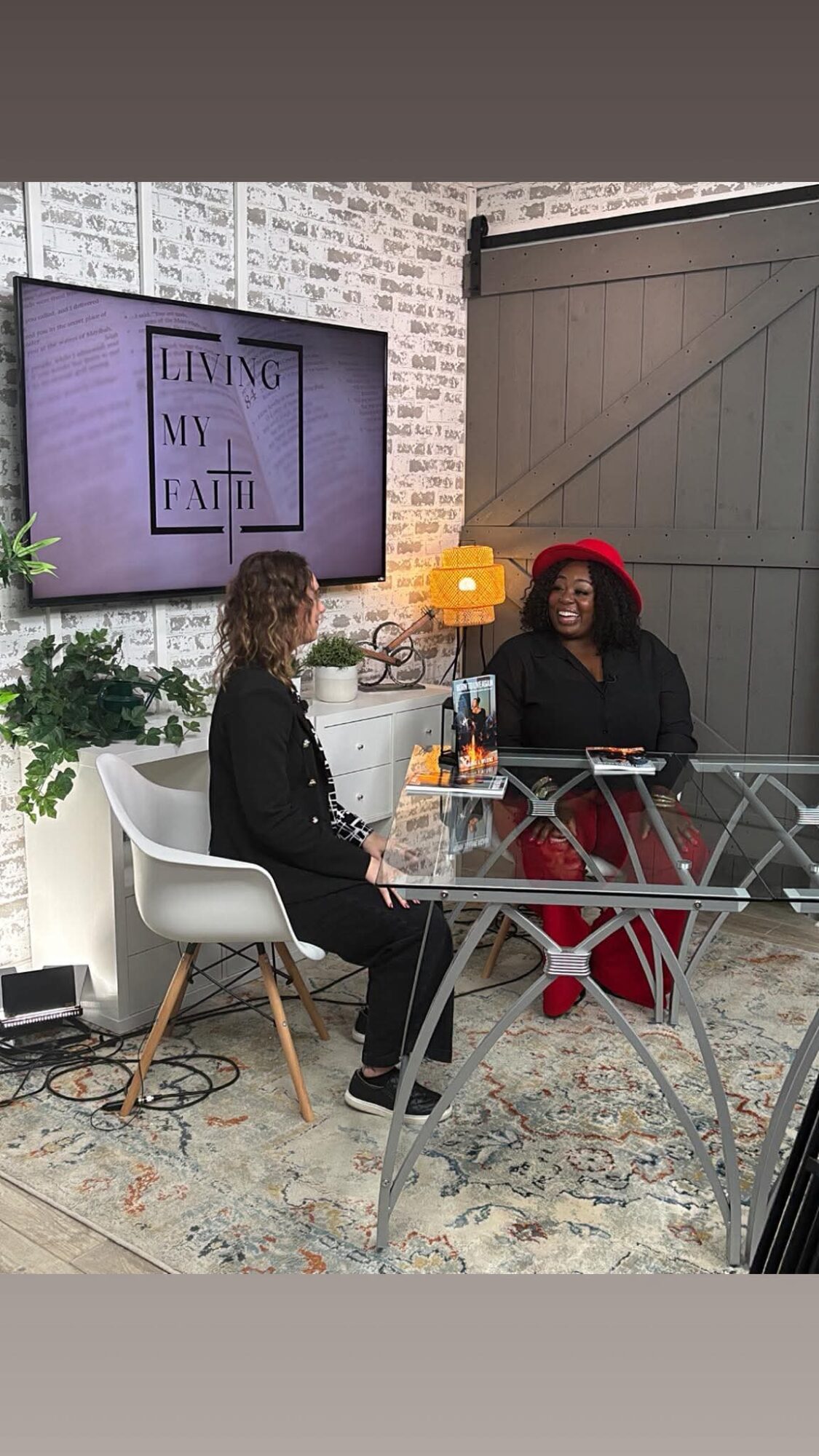Two women sit at a glass table in a room with a brick wall, a large screen, and a lamp, engaged in conversation.