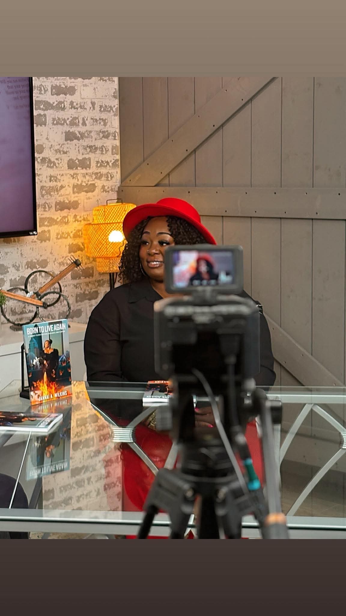 Person wearing a red hat and black outfit sitting at a table with a camera in front, in a cozy indoor setting.
