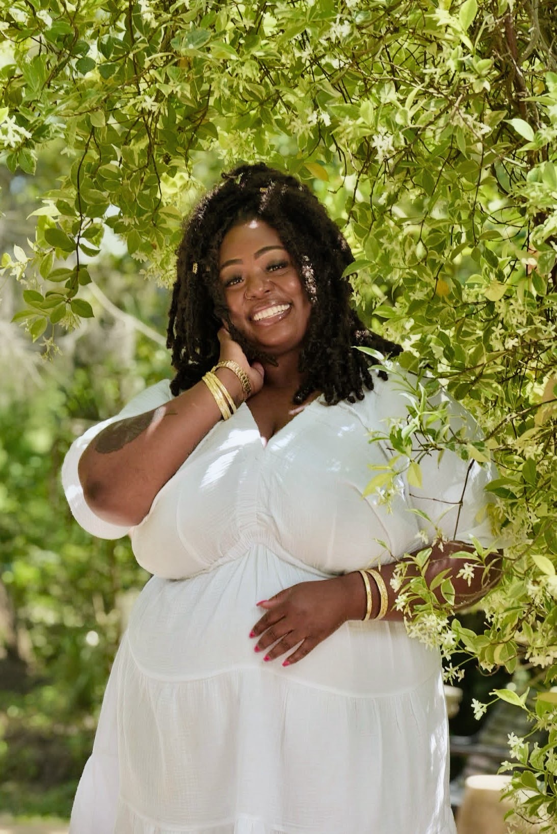 Smiling woman with curly hair in a white dress, standing outdoors among green leaves.