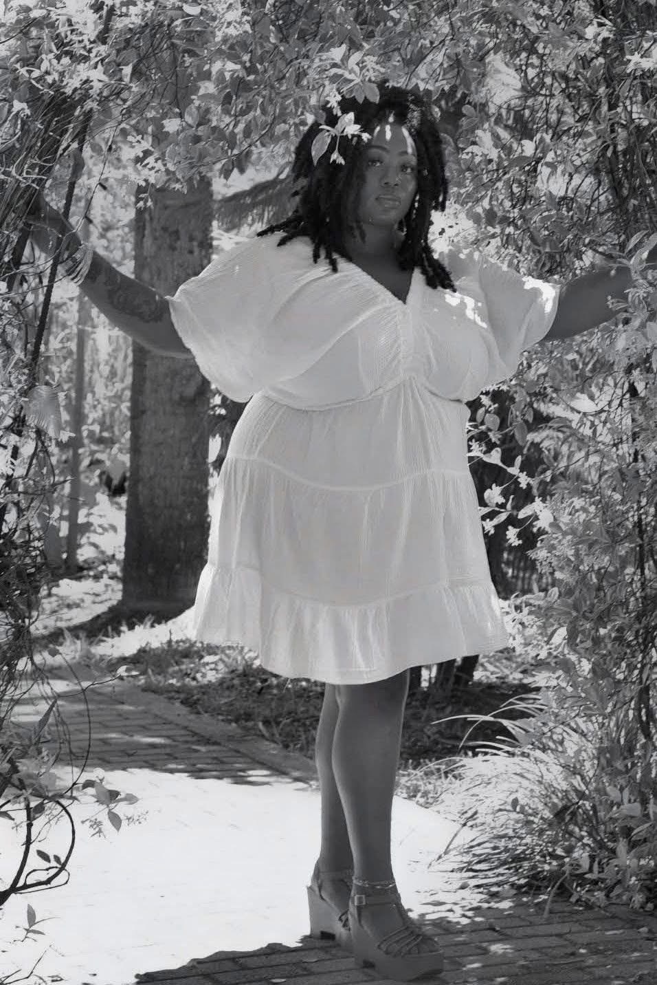 Person standing outdoors with arms outstretched, wearing a white dress, surrounded by trees and foliage