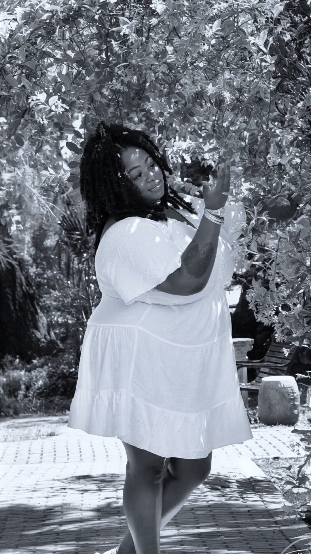 Person with braided hair in a white dress standing outdoors among trees and plants, holding a phone.