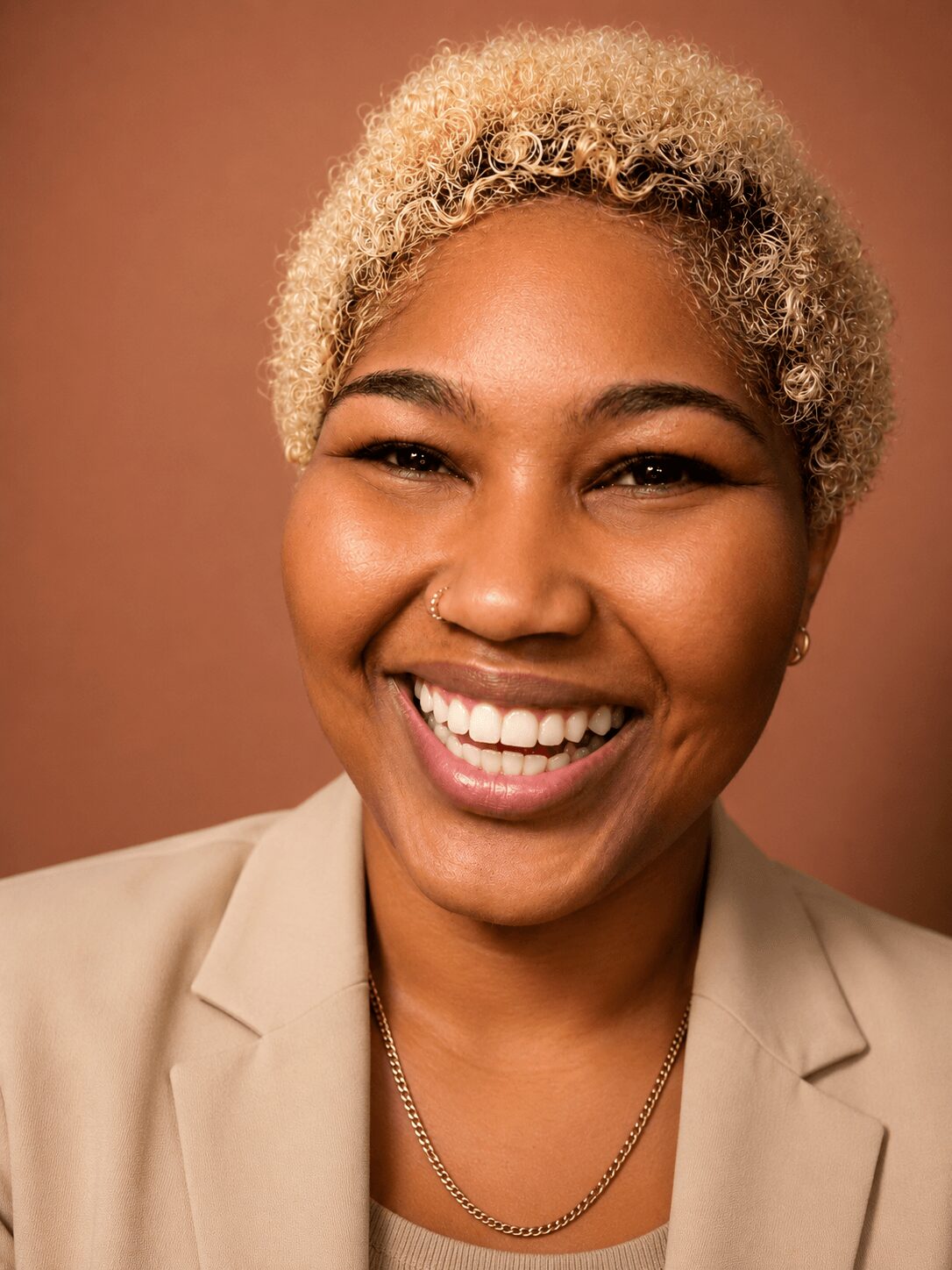 Smiling woman with short, curly blonde hair, wearing a beige blazer and a necklace, against a brown background.