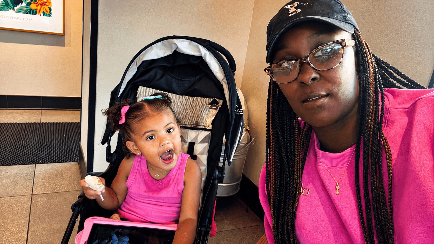 Two females, one young girl in a pink top and a woman with glasses and braids, sitting indoors near a stroller.