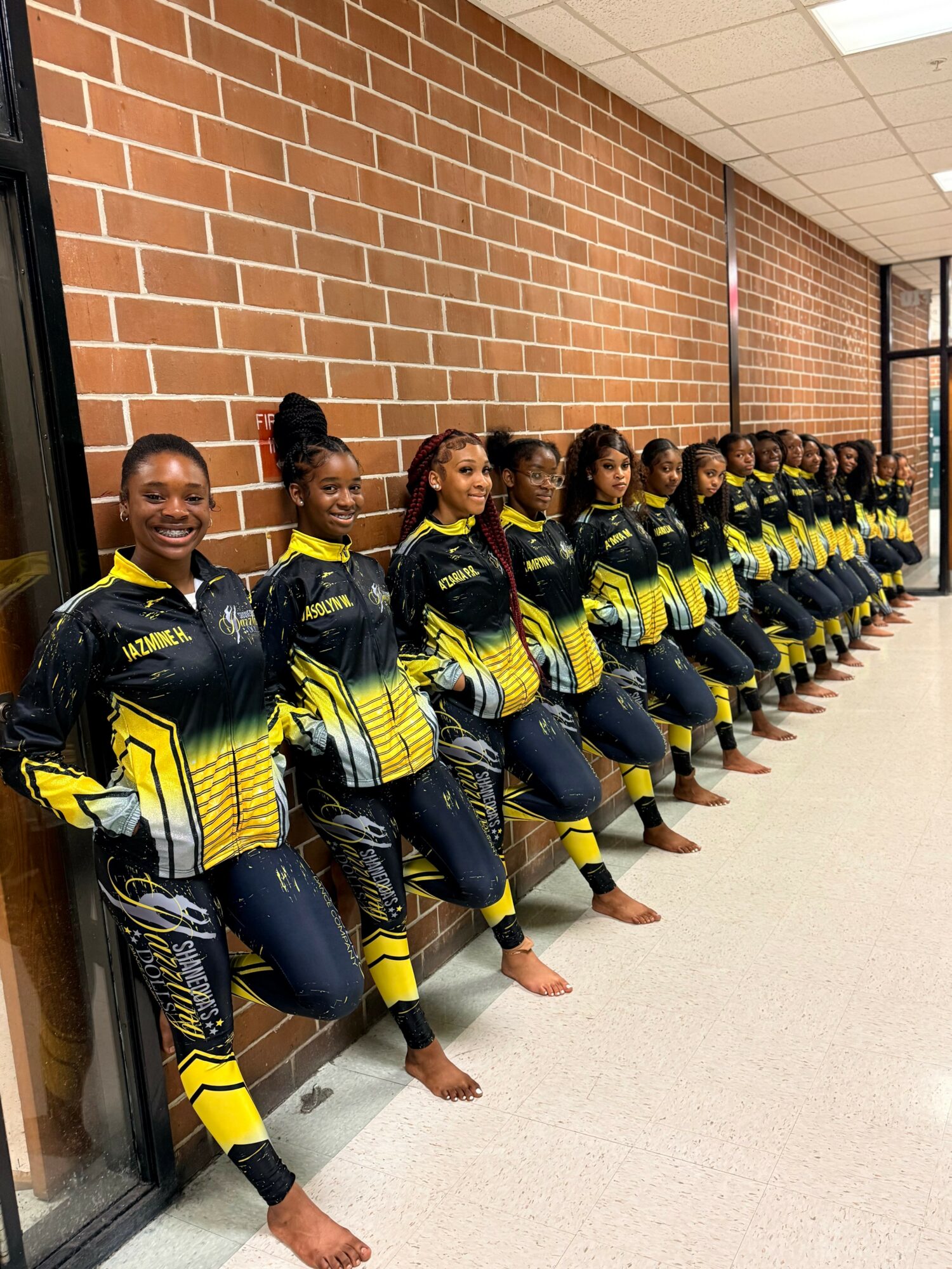 Group of young women in matching sports uniforms standing against a brick wall, smiling, with some barefoot, in a hallway.