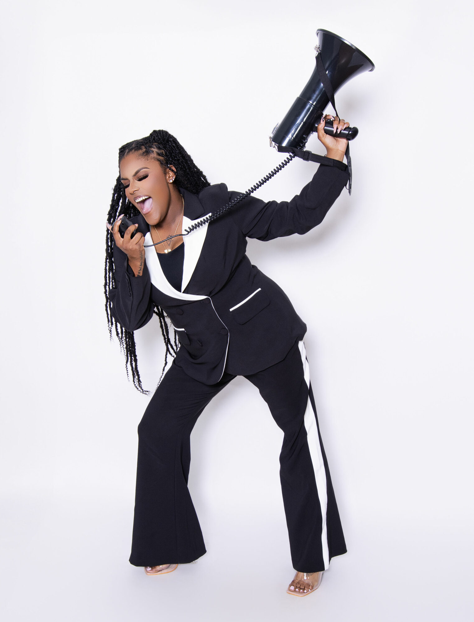 Woman in black suit holding a megaphone and talking on a phone, smiling, against a white background.