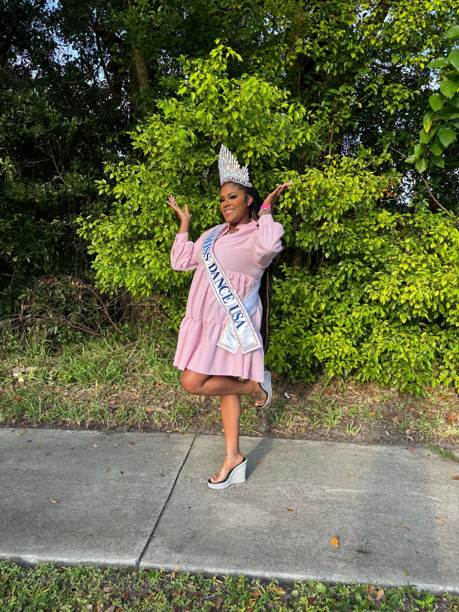 Person in pink dress and crown holding a sash, standing outdoors near green bushes and sidewalk.