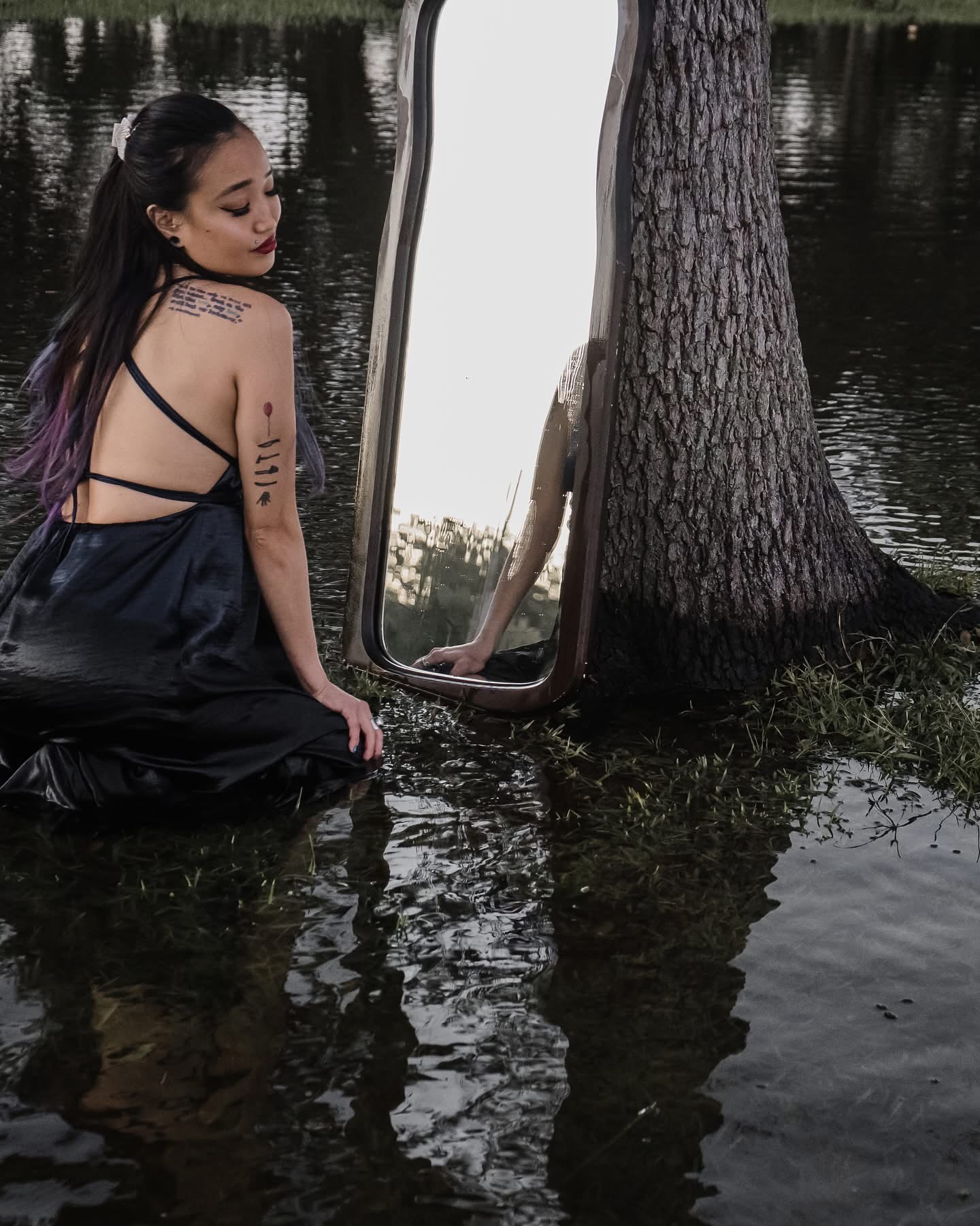 Woman with long dark hair and tattoos kneels by water, looking at a mirror reflecting a tree and water, outdoors.