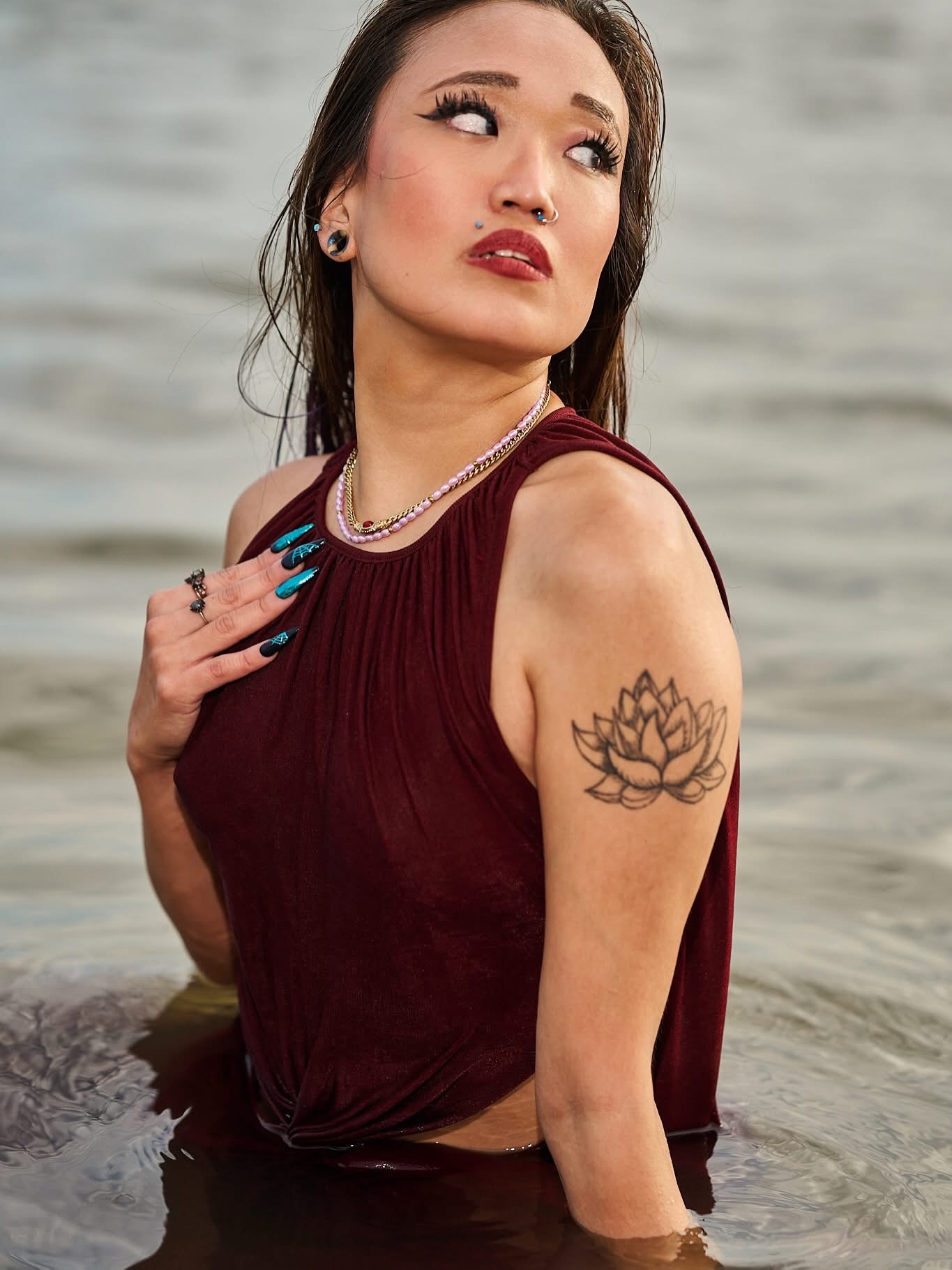 Woman with dark hair, makeup, and a tattoo on her upper arm, standing in water, wearing a sleeveless top and jewelry.