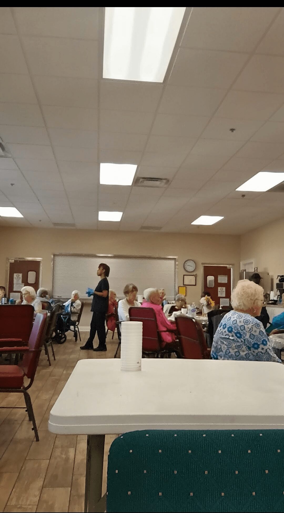 People seated at tables in a room with a ceiling light, some standing and talking, with a white table in foreground.