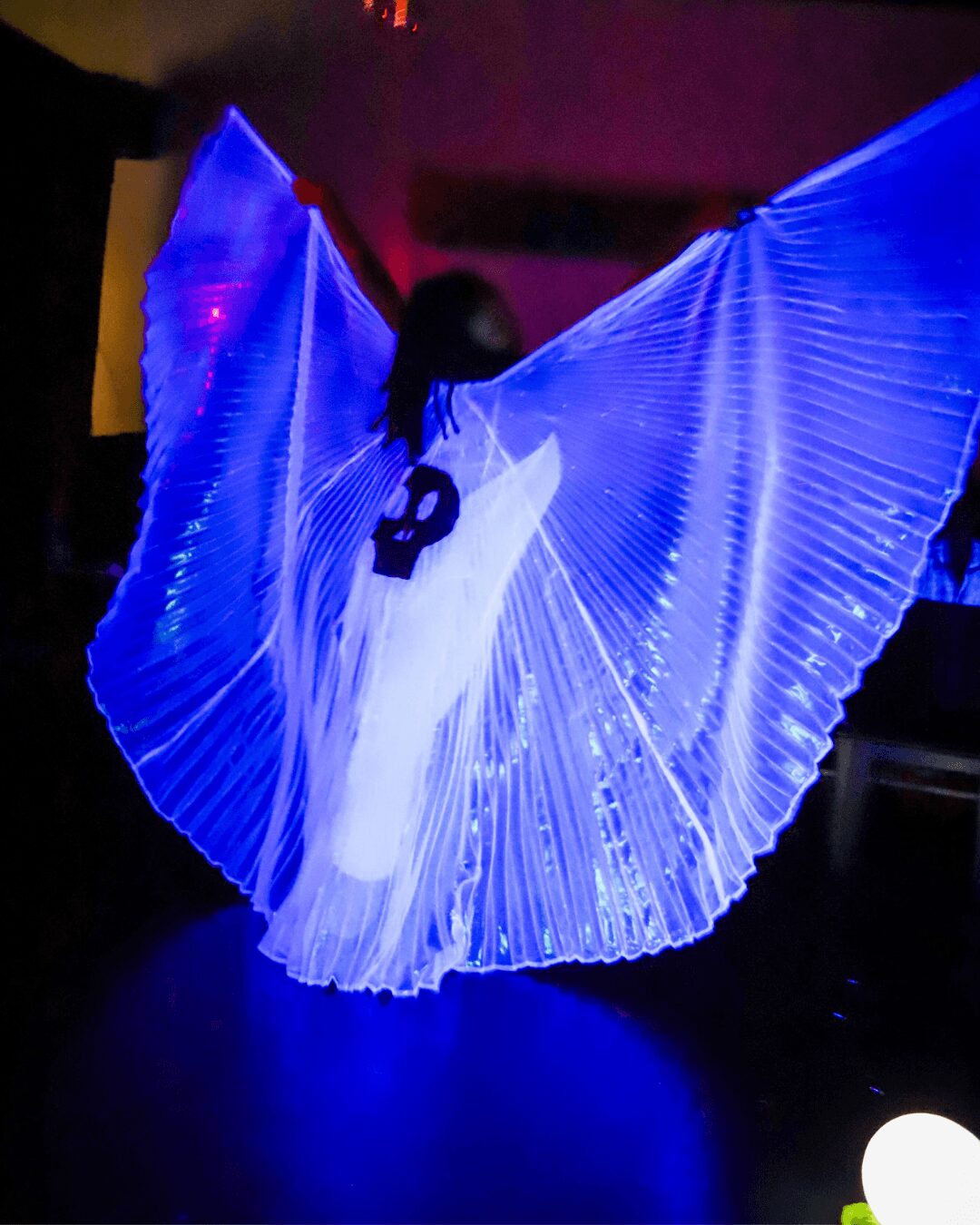 Person performing with large illuminated blue wings, dark background, colorful lighting, and a glowing object nearby.