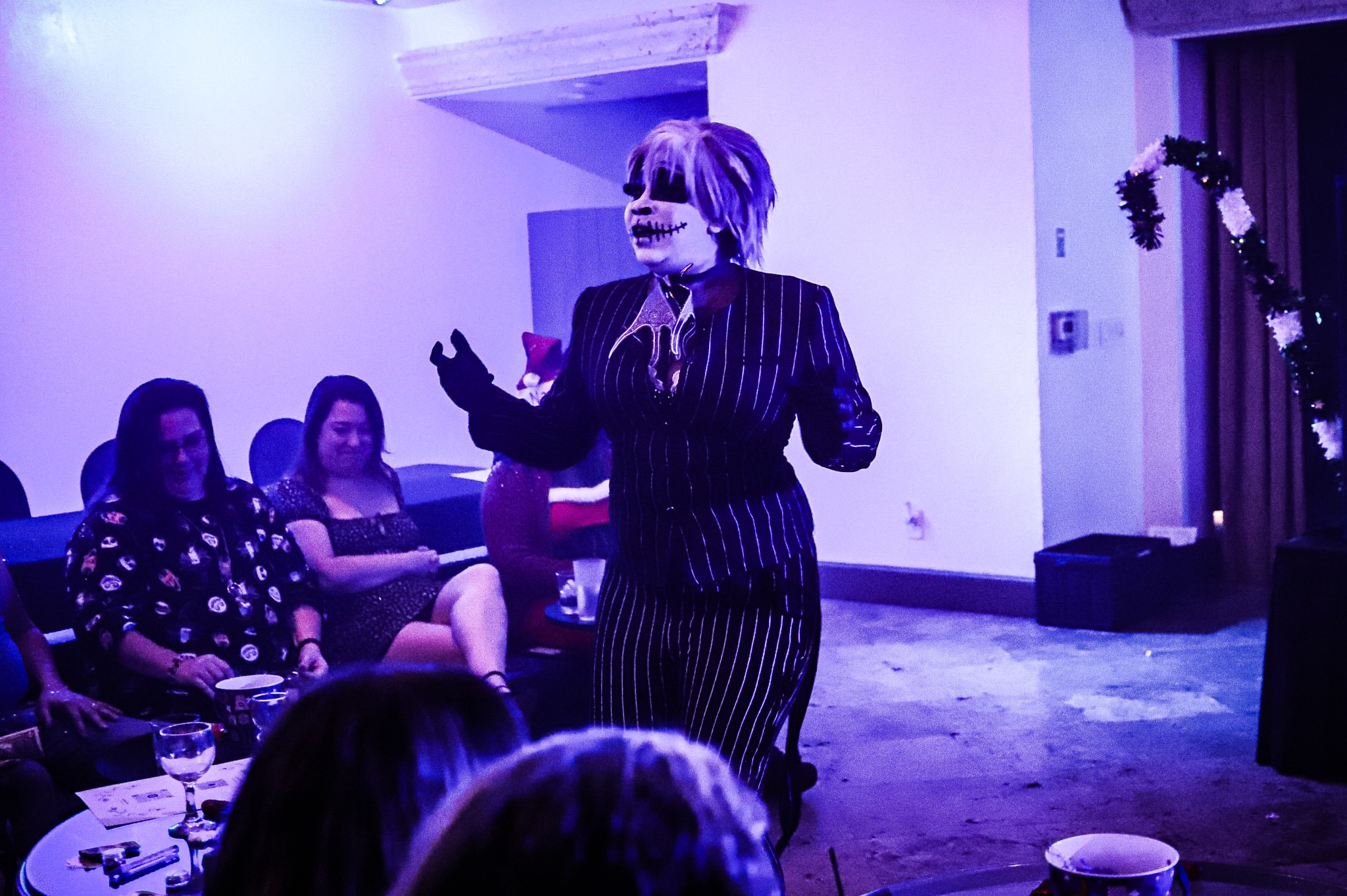 Person with face paint and striped suit speaking to an audience in a dimly lit room.