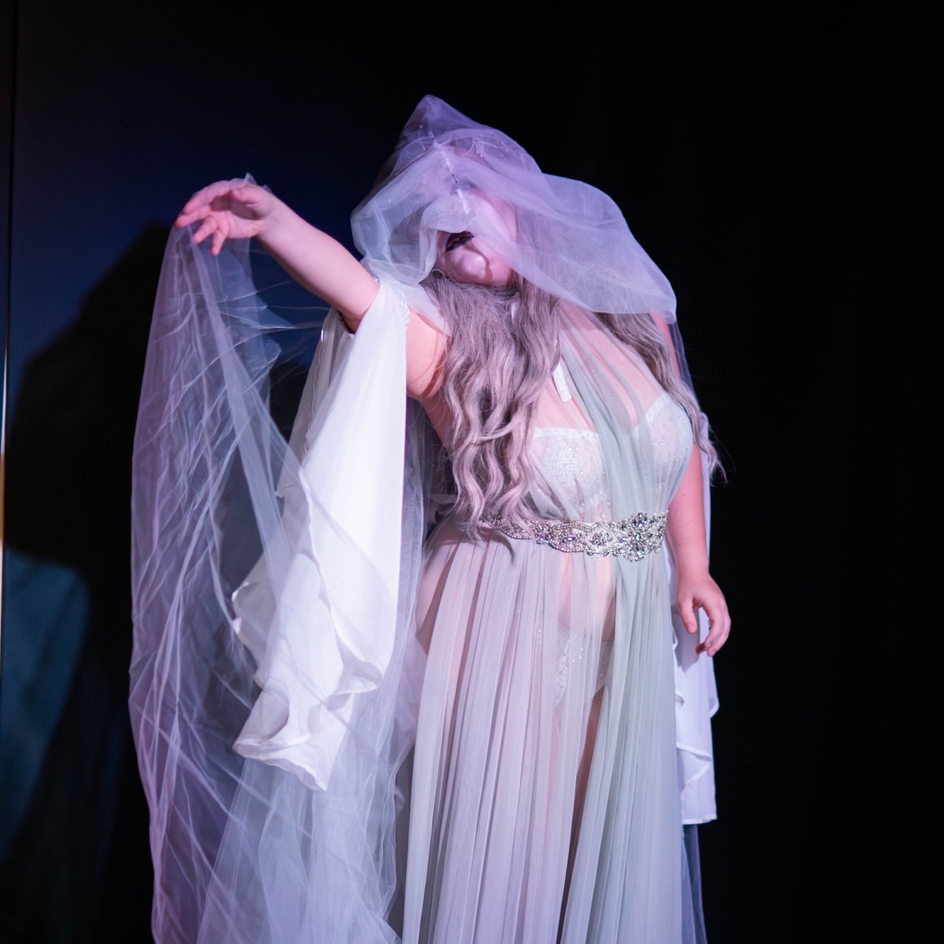 Person in flowing dress with veil covering face, posing with arm raised, against dark background.