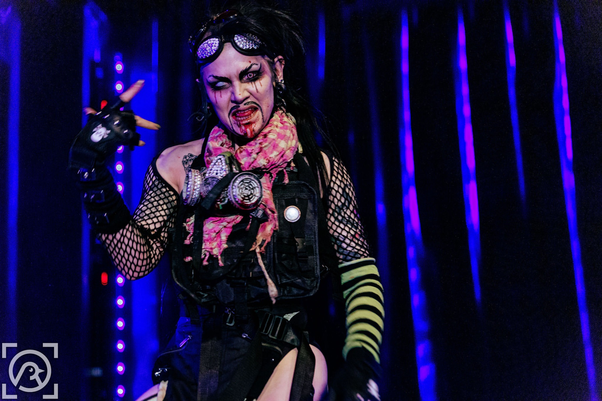 Performer with makeup, fishnet clothing, and colorful accessories on stage with blue and purple lights, expressive pose.