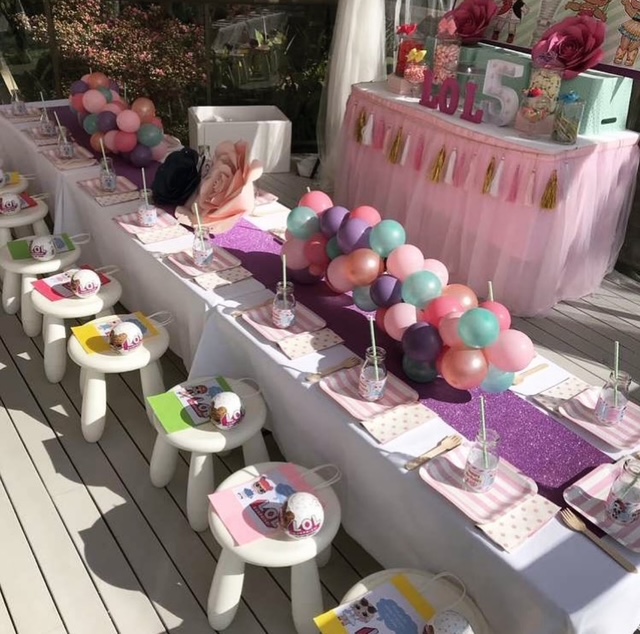 Party table decorated with balloons, cupcakes, and pink decorations, set outdoors with sunlight and wooden flooring.